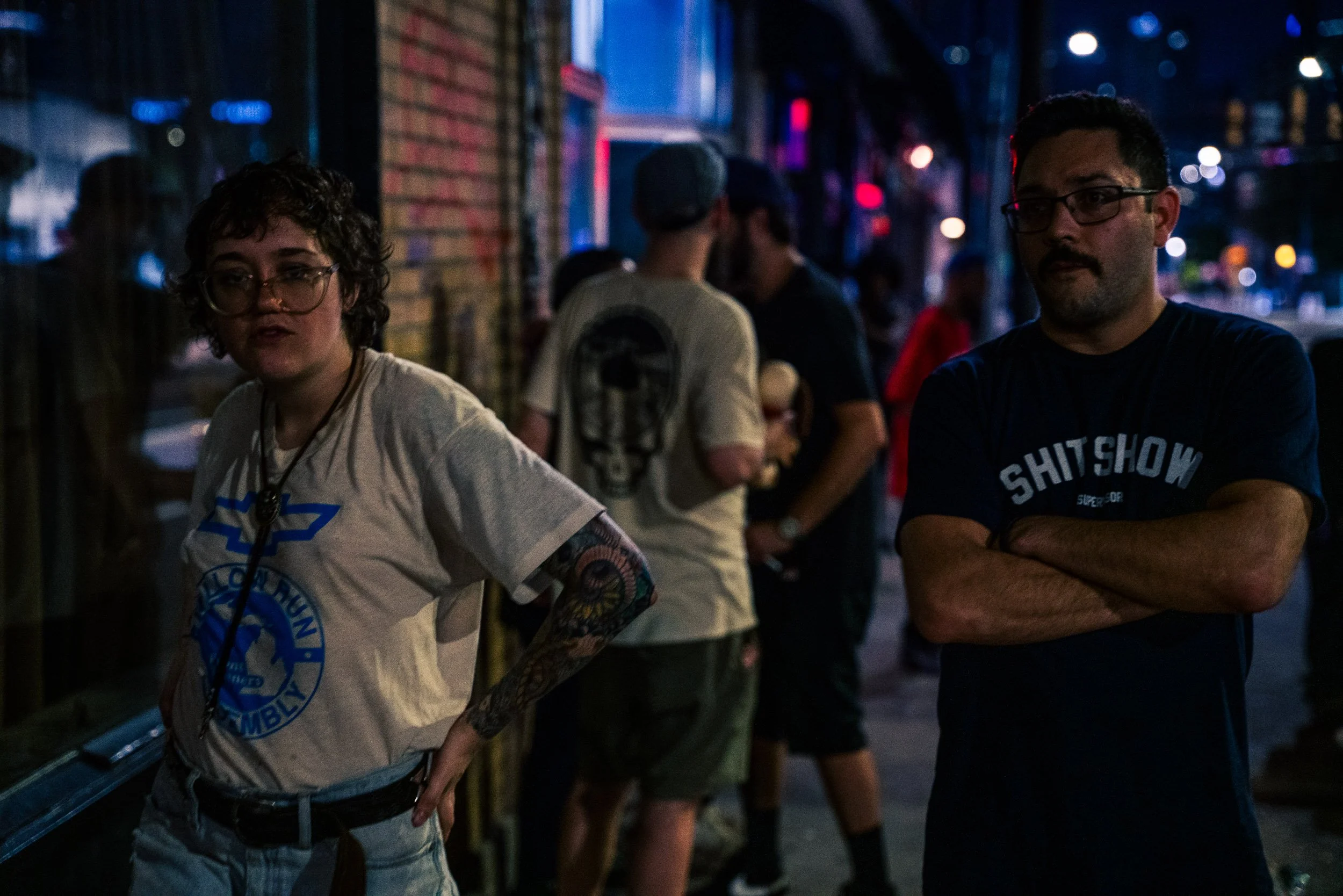 Two people, one with glasses and short curly hair wearing a white t-shirt with a blue logo, and the other with glasses and dark hair wearing a dark t-shirt, standing outdoors on a city street at night with other people and colorful lights in the back
