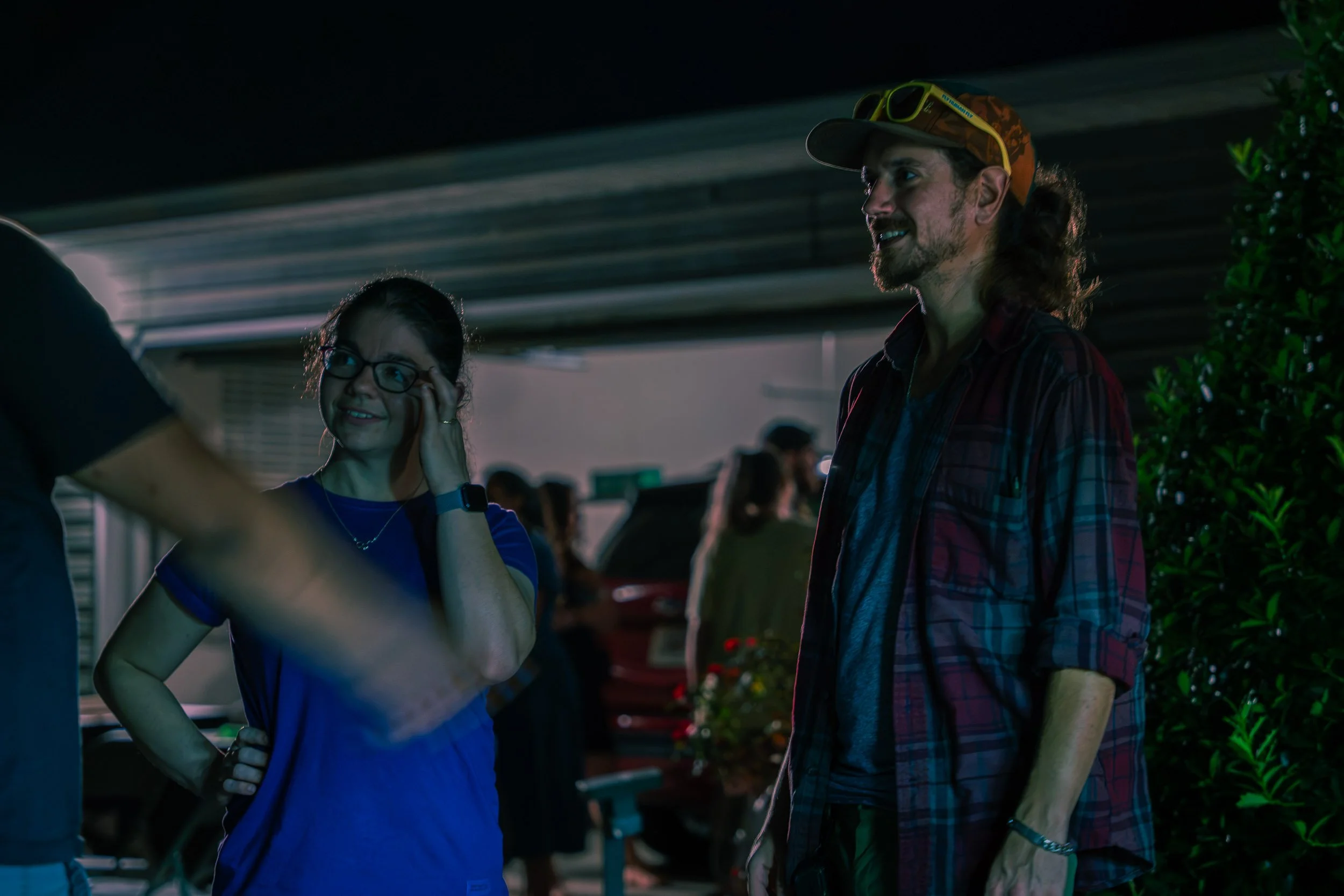 A woman and a man are engaged in a conversation at night, standing outdoors near some greenery and parked cars.