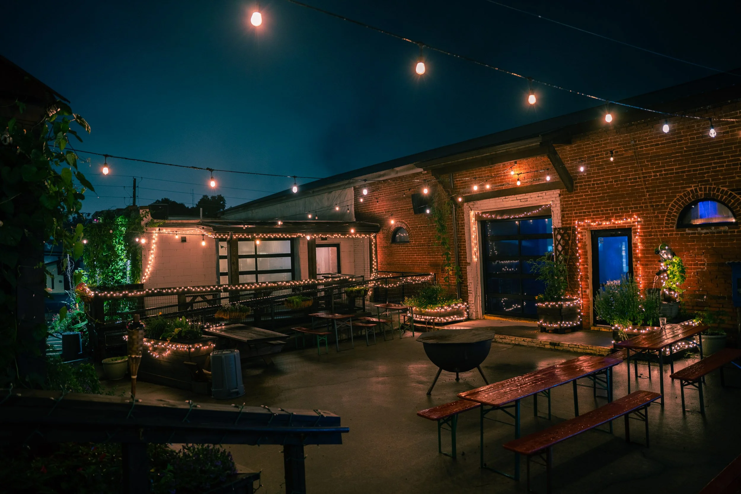 Nighttime outdoor patio with string lights, wooden tables and benches, red brick building, and a clear, dark sky.