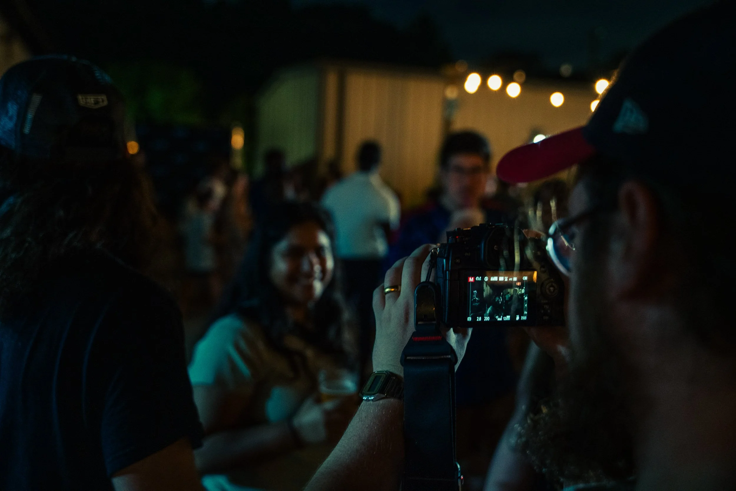 Nighttime scene of a group of people gathered outdoors, some wearing hats, with string lights in the background. A person in foreground is taking a photo with a DSLR camera.
