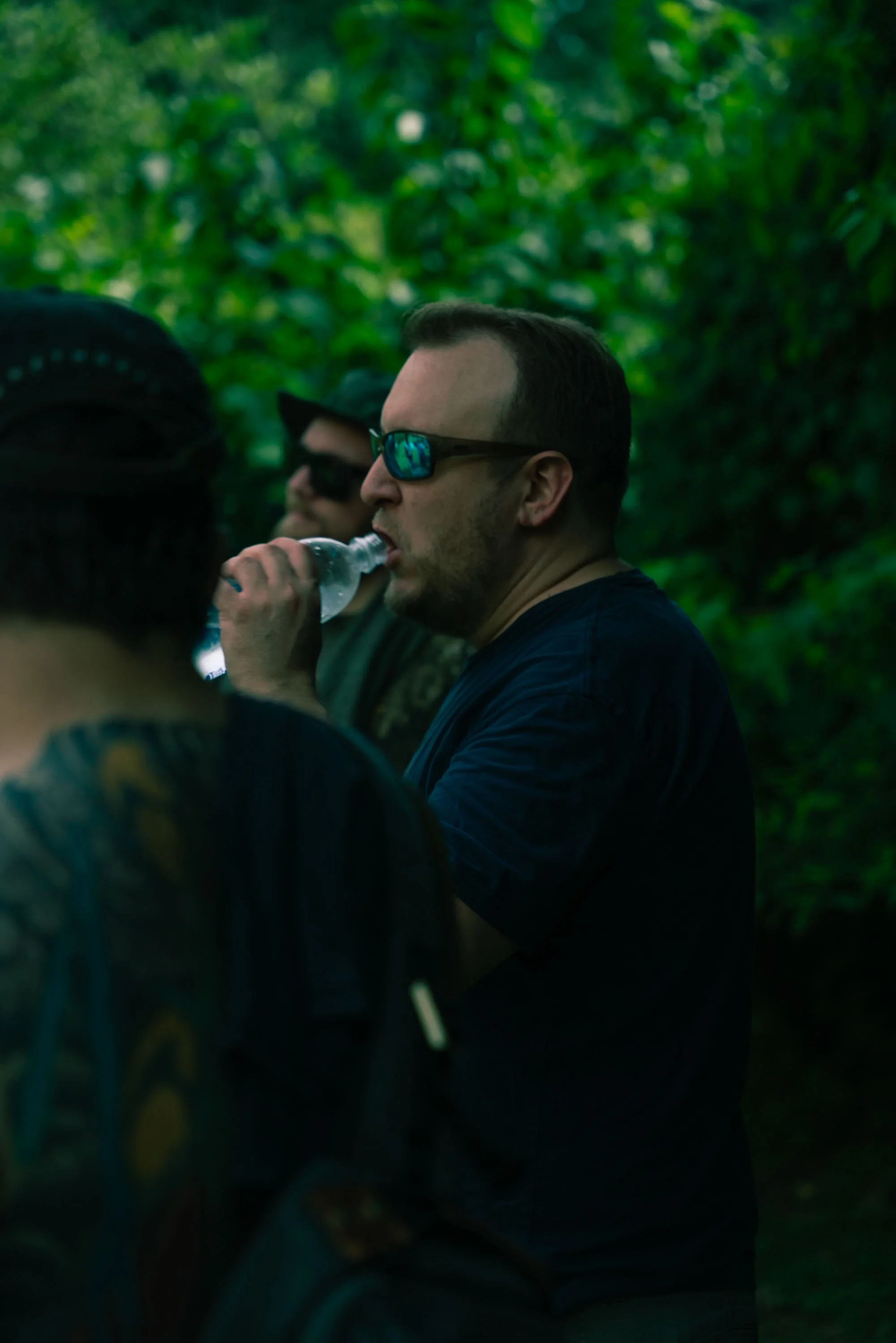 Three men standing outdoors in a wooded area, one drinking water from a plastic bottle, two wearing sunglasses, surrounded by green foliage.