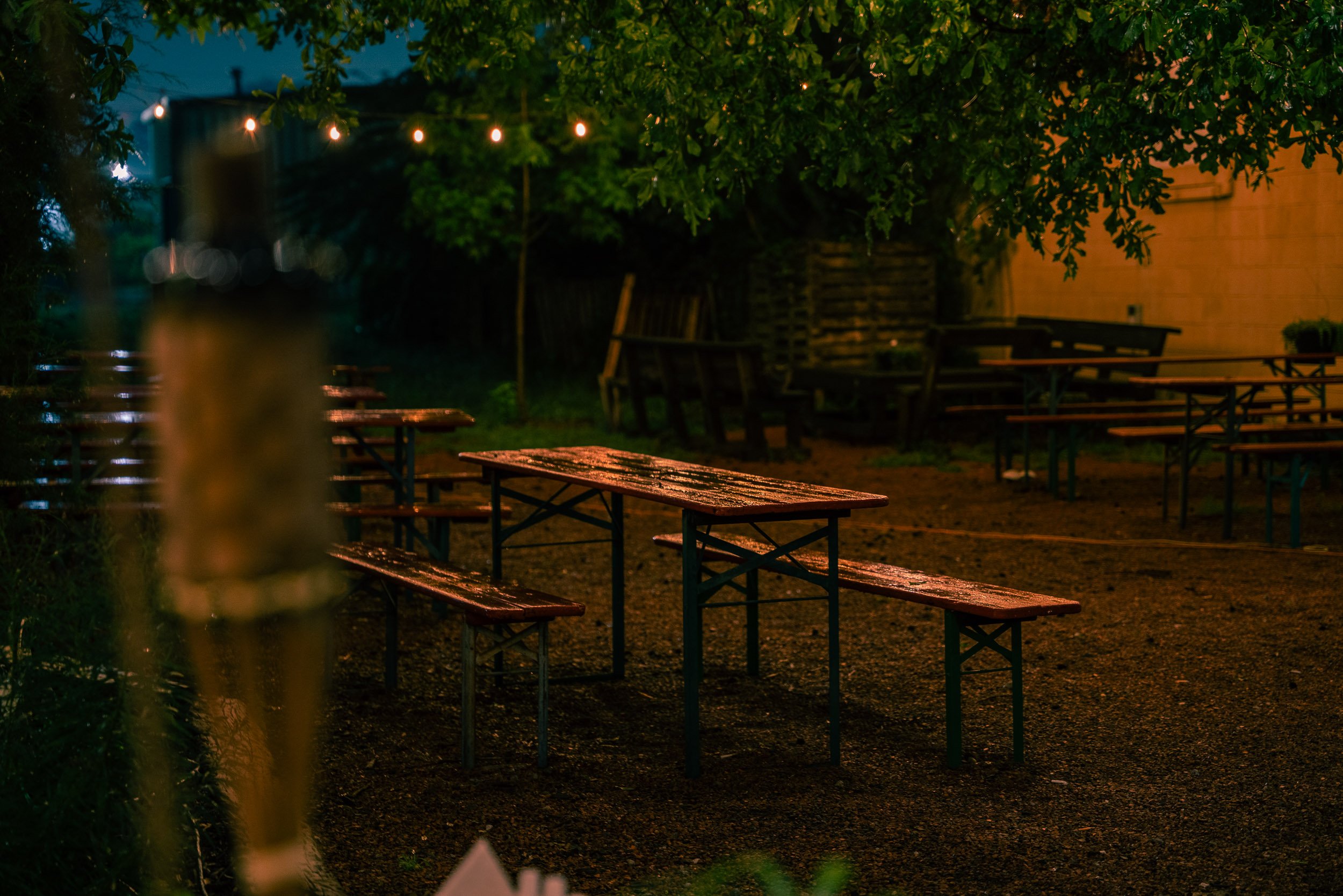 Empty outdoor seating area with wet tables and benches, surrounded by trees and string lights at dusk or night.