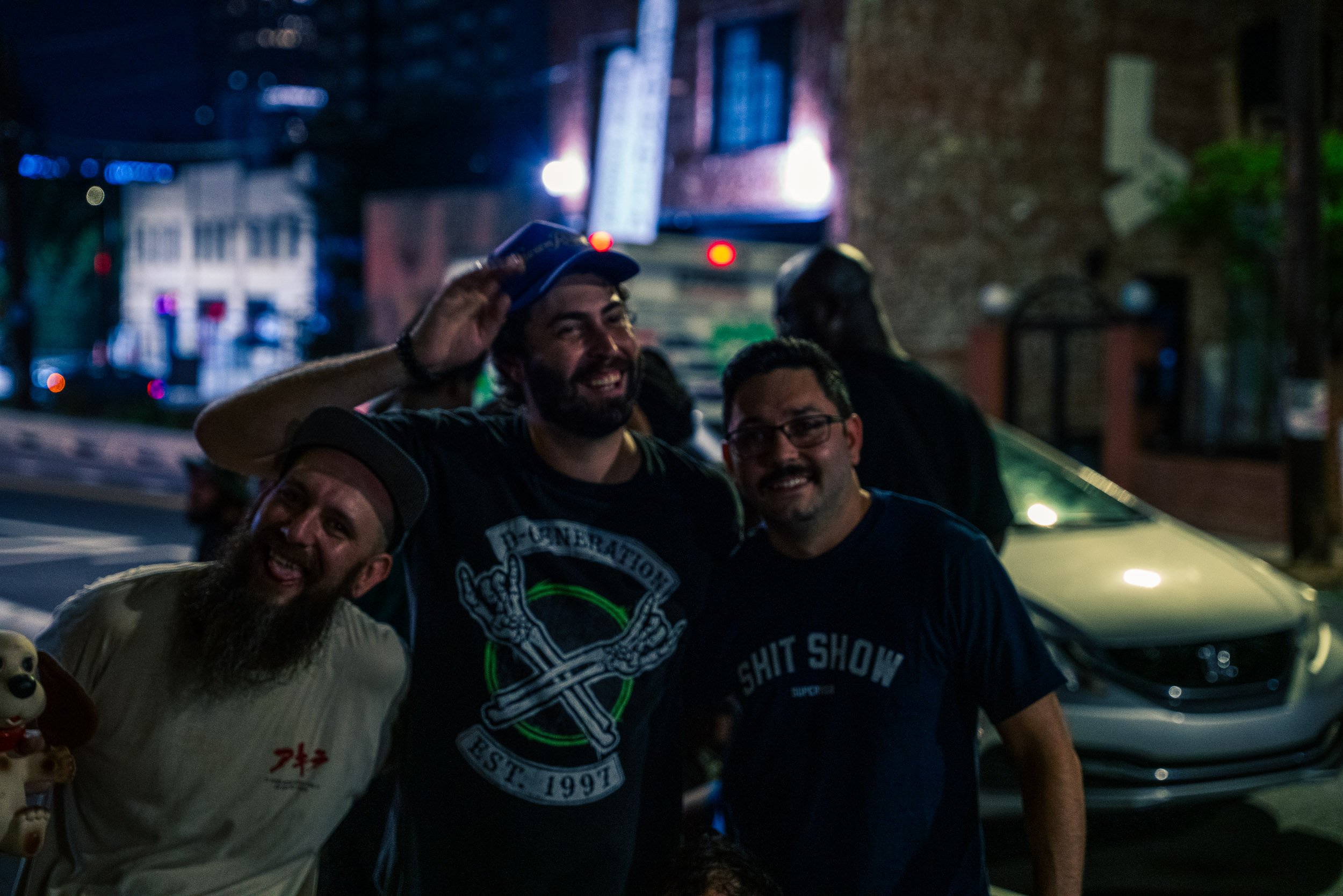 A group of smiling men posing together on a city street at night, with blurry lights and buildings in the background.