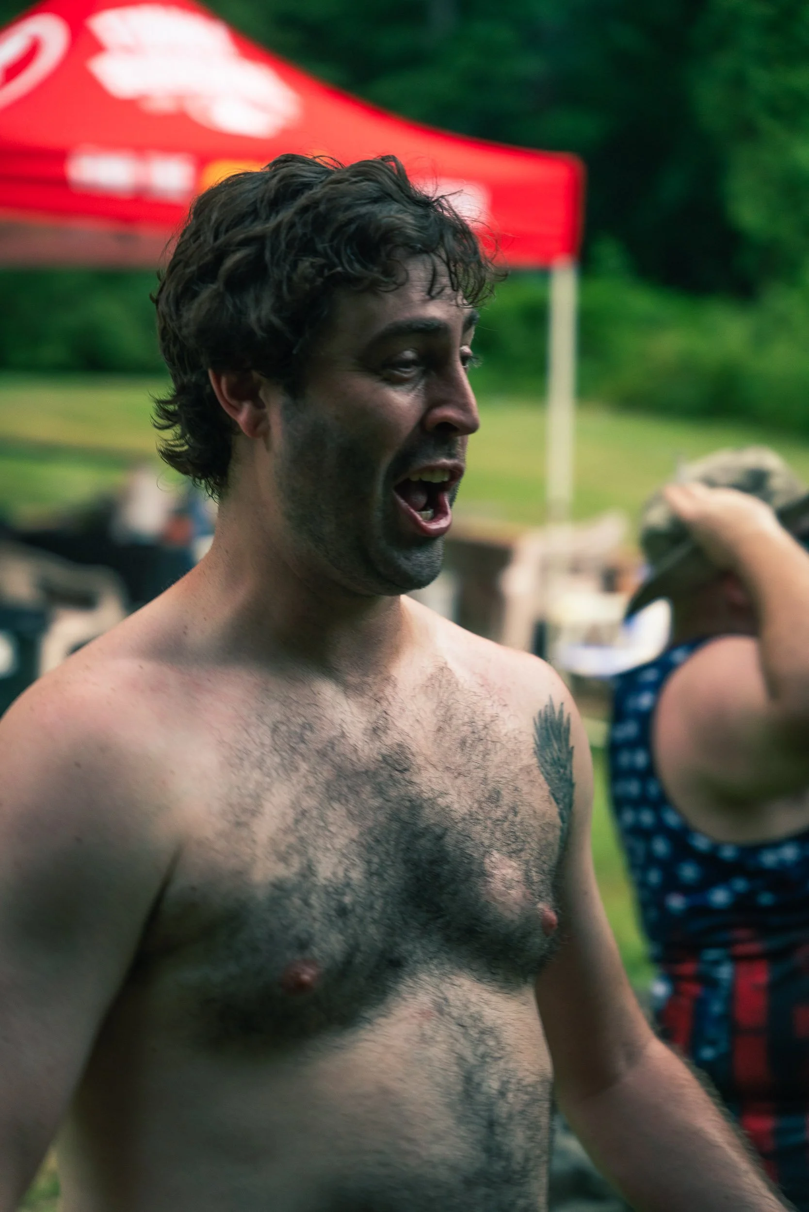 A shirtless man with curly dark hair and tattoos on his chest, standing outdoors during the daytime, with a red tent and green trees in the background.