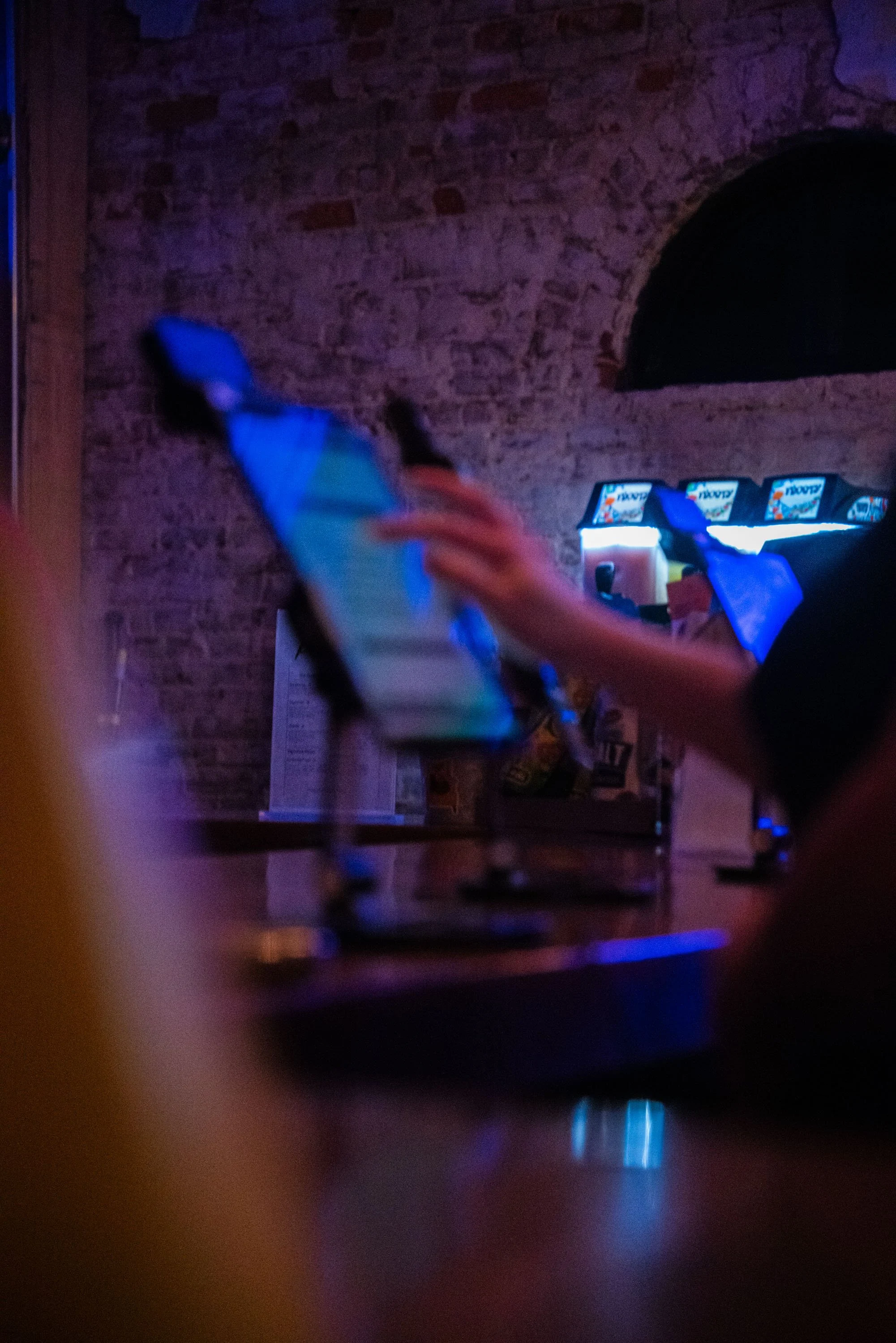 Blurred image of a person's hand holding a phone in a dimly lit setting with a brick wall in the background and arcade game machines in the distance.