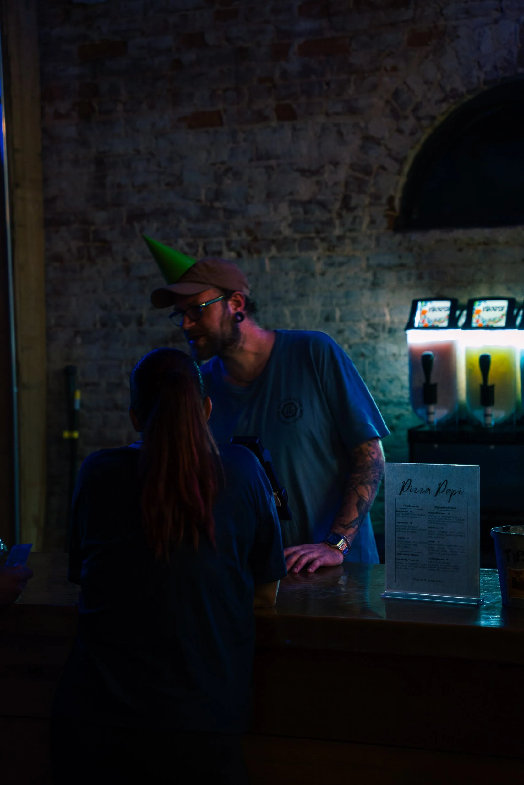 A man with glasses, tattoos, and a watch wearing a party hat is at a bar or restaurant, talking to a woman with red hair. The background features a brick wall and a draft beer menu.