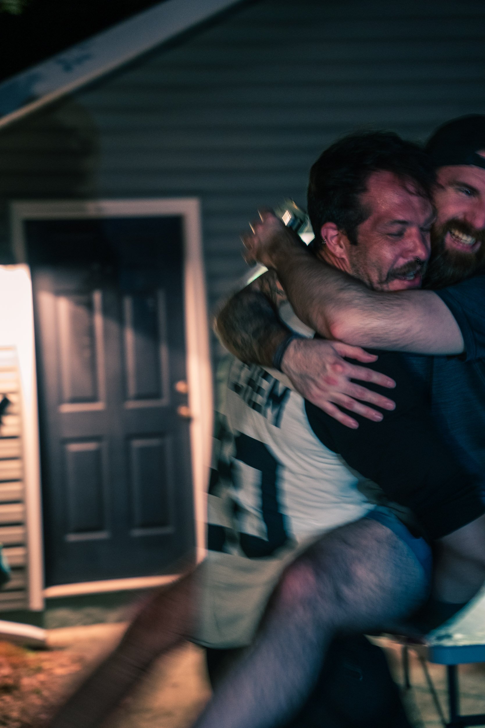 Two men hugging and smiling inside a room, with a computer monitor and a dark wall in the background.