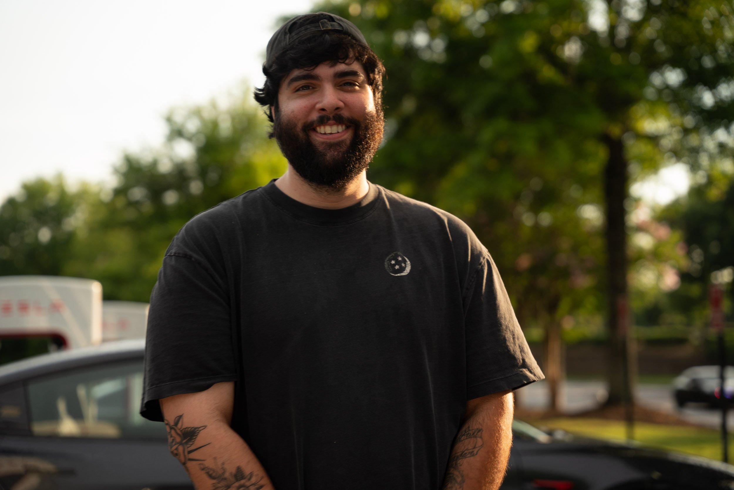 A young man with dark hair, beard, and tattoos, smiling outdoors in front of a blurred background of trees, parked cars, and a bright sky.