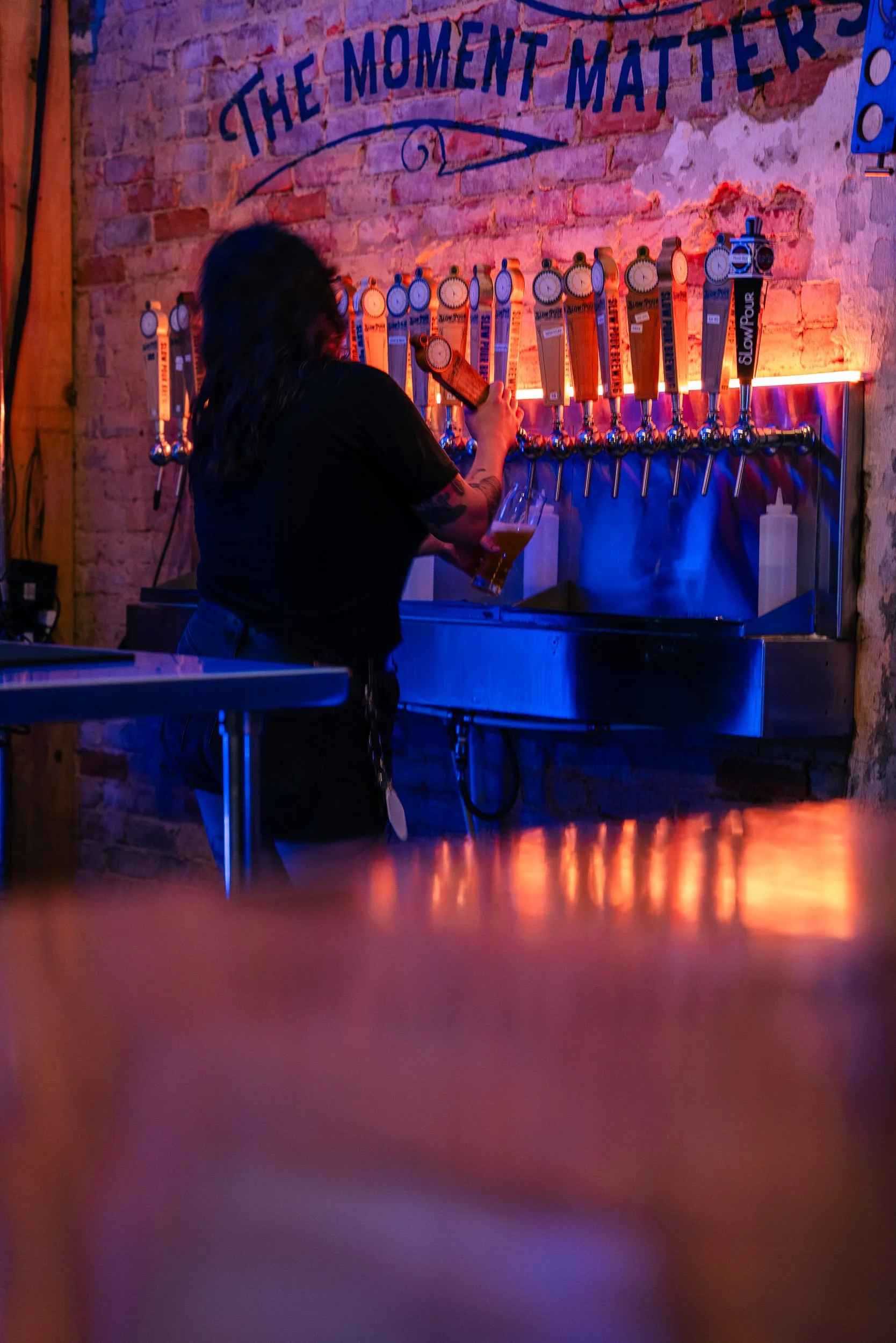 A woman with dark hair, dressed in black, standing at a bar with multiple beer taps, holding a glass of beer, with a sign on the wall that reads 'The Moment Matters'.