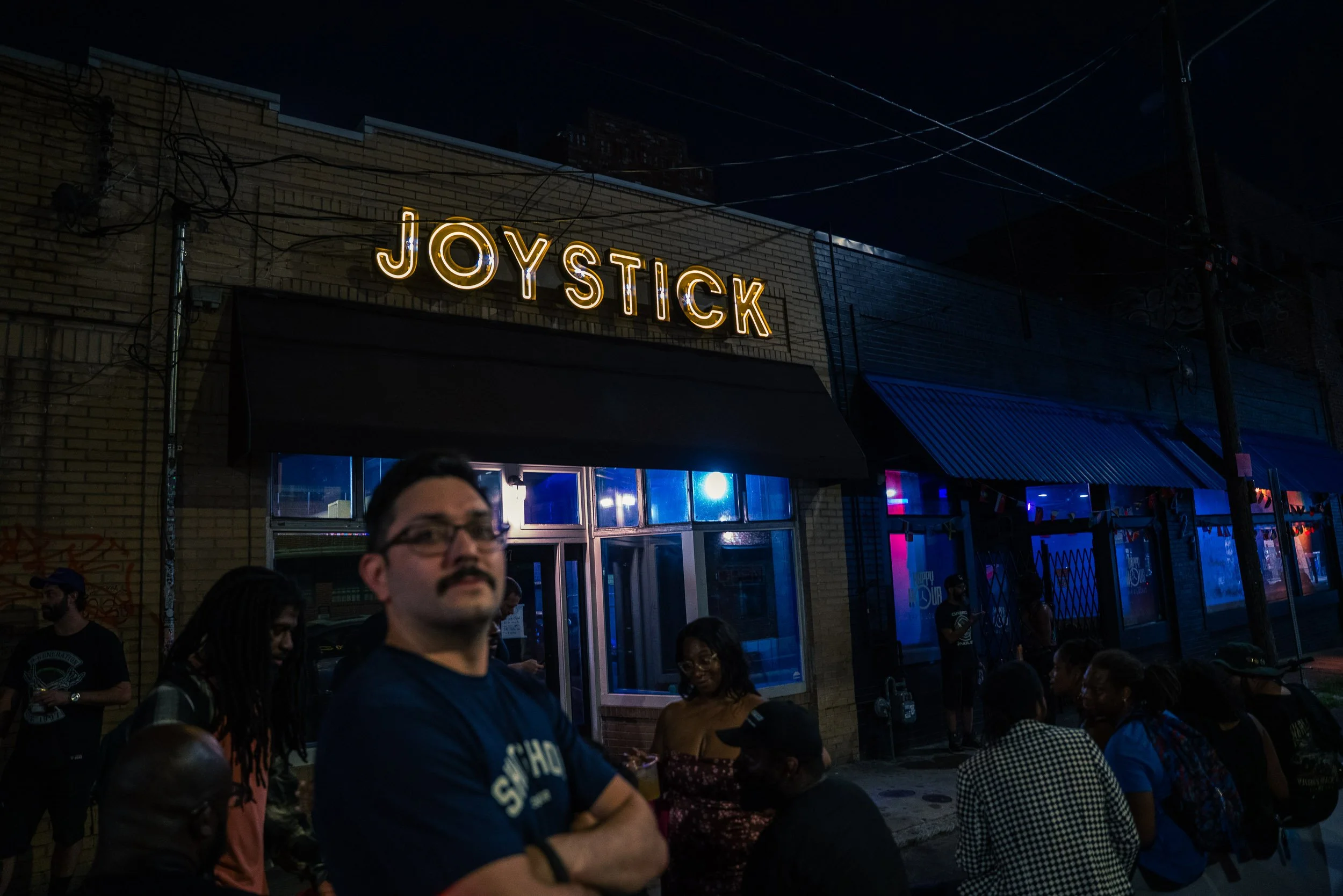 Night scene outside a bar or music venue with a bright neon sign that reads 'JOYSTICK'. Several people are gathered outside, some talking and others looking around. The building has yellow brick walls, and there are some power lines overhead.