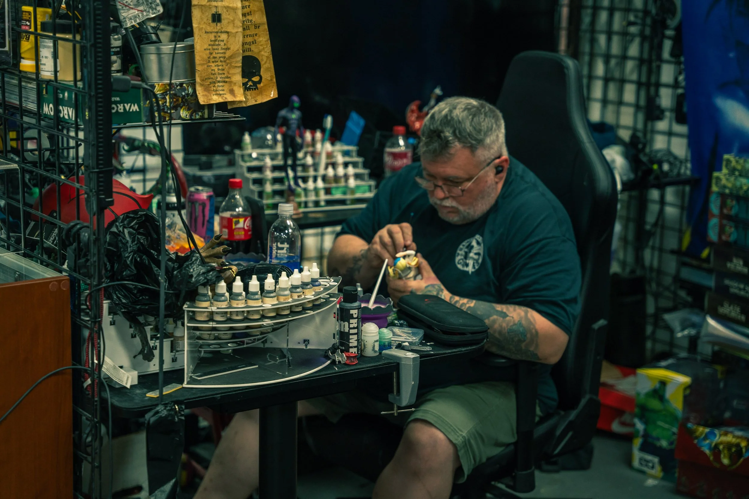 An older man with tattoos, glasses, and gray hair is sitting at a cluttered workbench, painting miniature figures. The workspace is filled with paints, brushes, and various supplies, with shelves and containers around him.