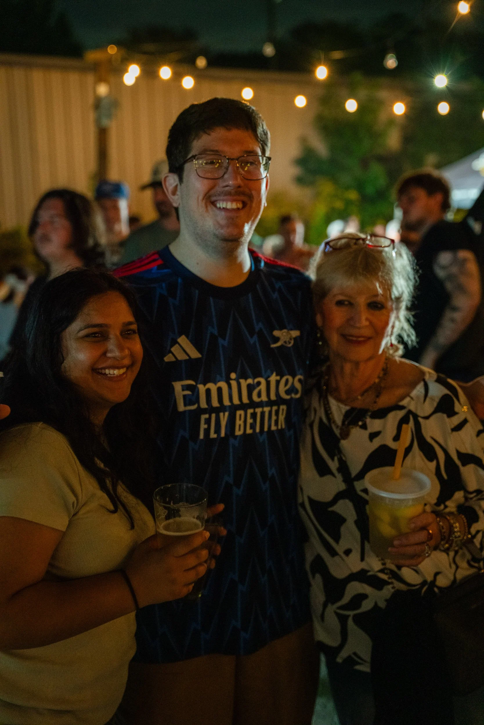 People at a nighttime outdoor gathering, smiling and holding drinks under string lights, with a wooden fence and trees in the background.