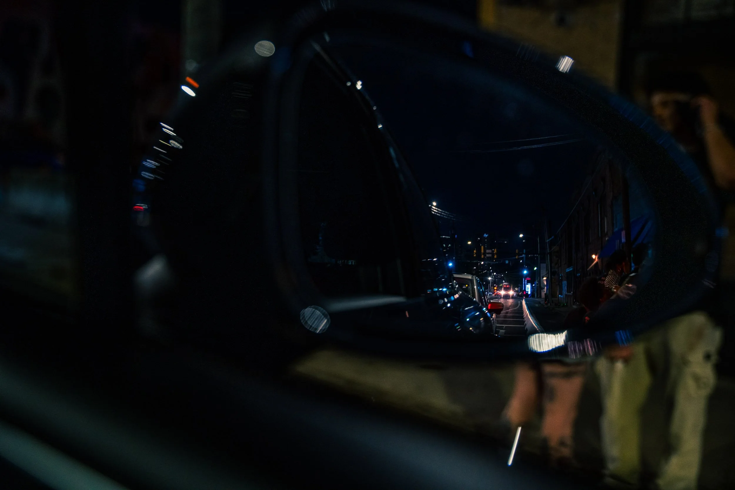 A nighttime street scene viewed through a vehicle's windshield, with cars parked along the side of the street and some cars driving in the distance, illuminated by streetlights and headlights.