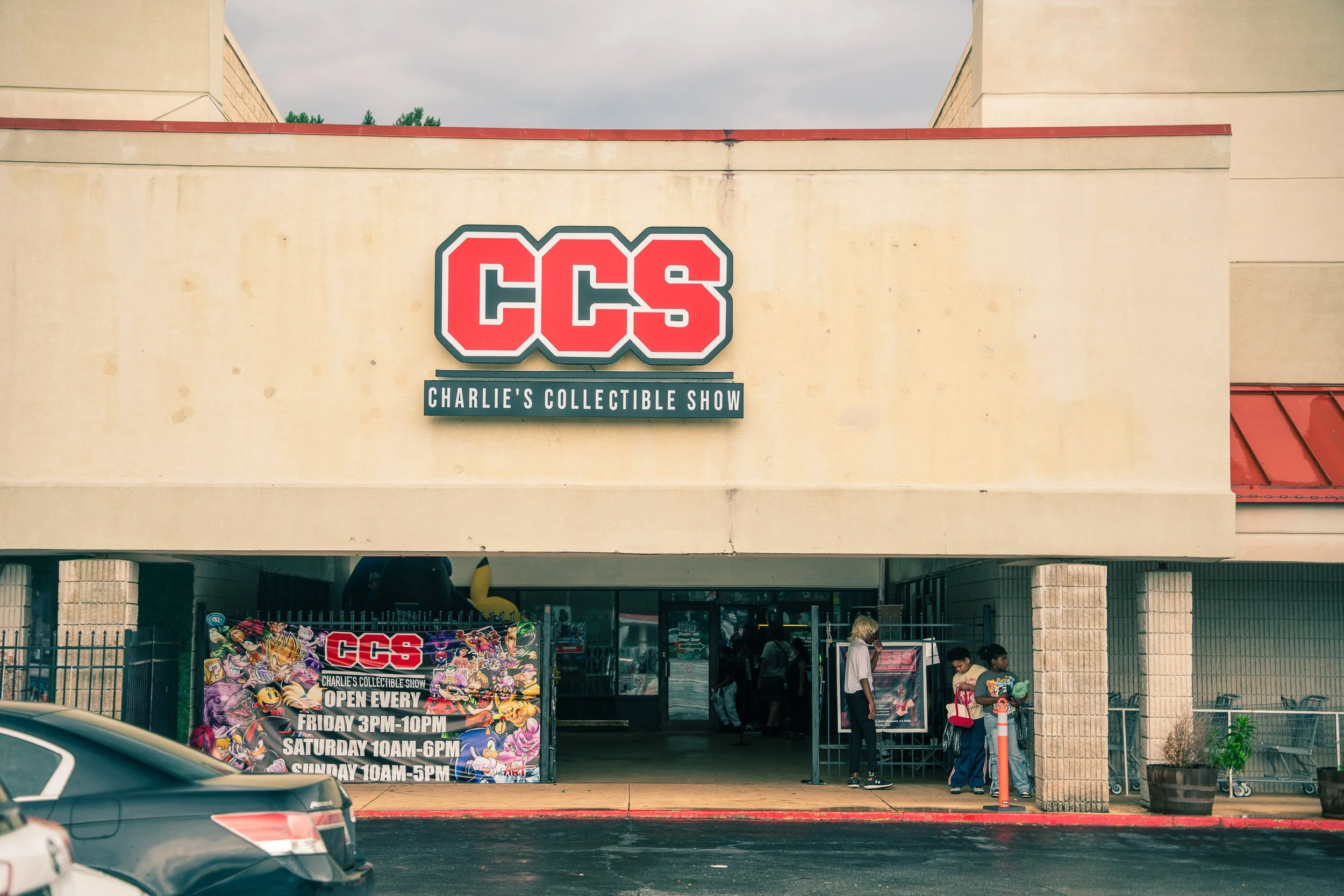 The entrance to Charlie's Collectible Show (CCS) with a large sign and a promotional poster indicating the show is open every Friday to Sunday. Several people are gathered near the entrance, and cars are parked in front.