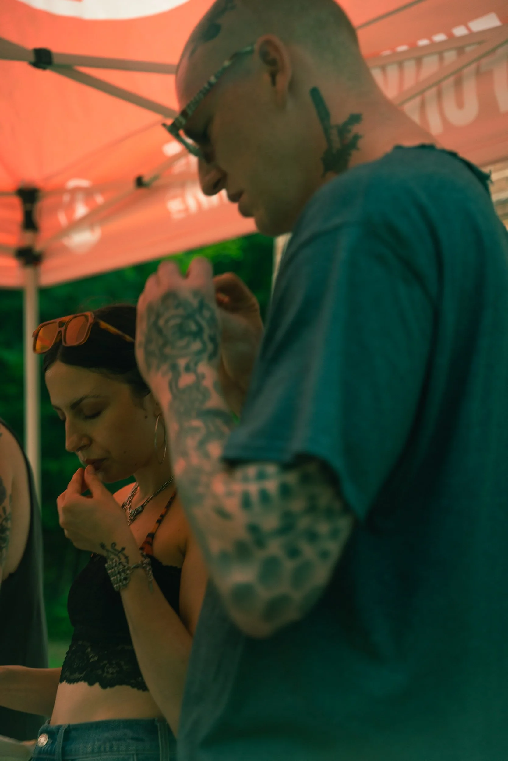 A close-up of a man with tattoos and glasses talking to a woman with sunglasses on her head and wearing a black lace top, under an orange canopy.