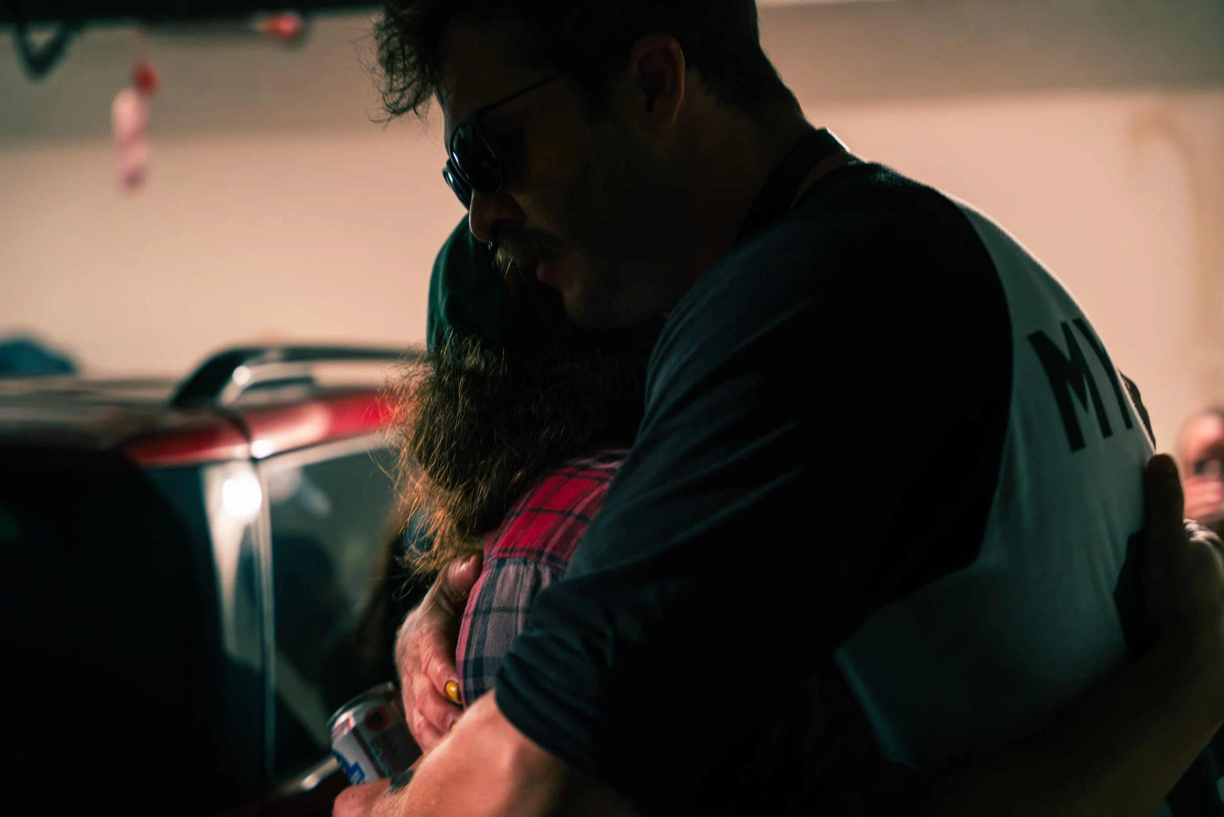 A man wearing sunglasses and a black shirt, hugging a person in a red plaid shirt. They are indoors with a dimly lit background.