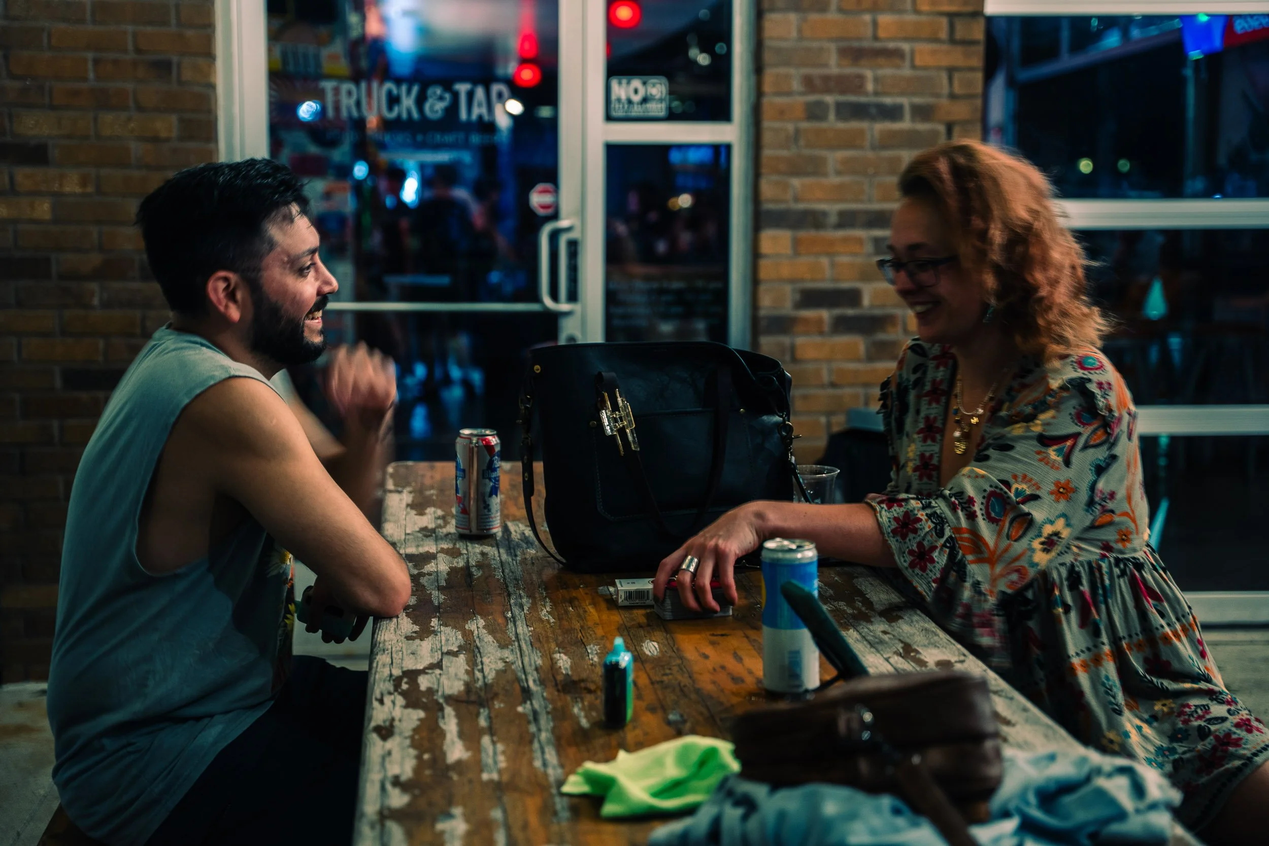 Two people, a man with dark hair and beard wearing a sleeveless shirt and a woman with red hair and glasses wearing a floral blouse, are sitting at a rustic wooden table inside a dimly lit restaurant or bar, smiling and engaging in conversation, with