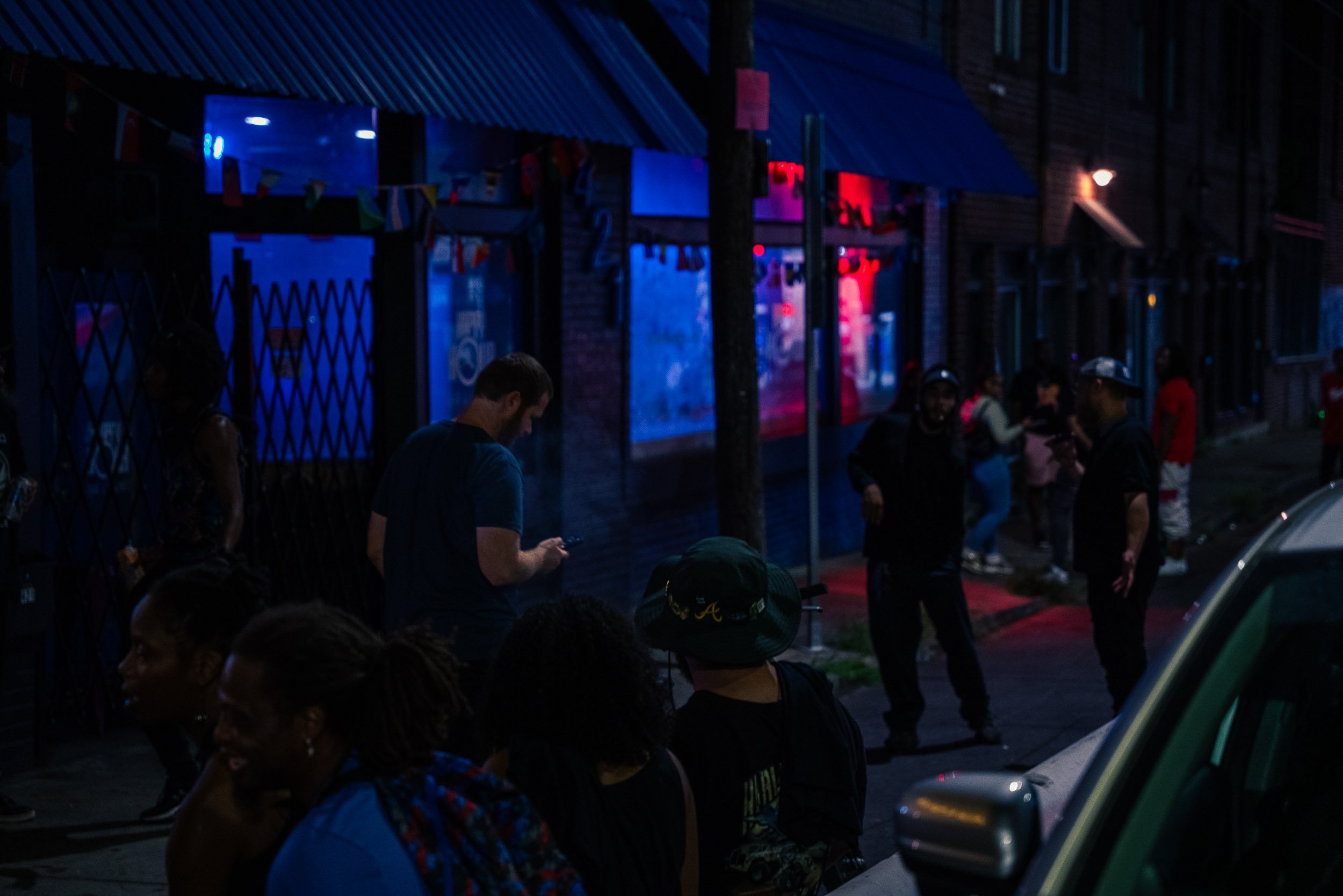 People standing on sidewalk at night in front of storefront with neon signs, some looking at phones.