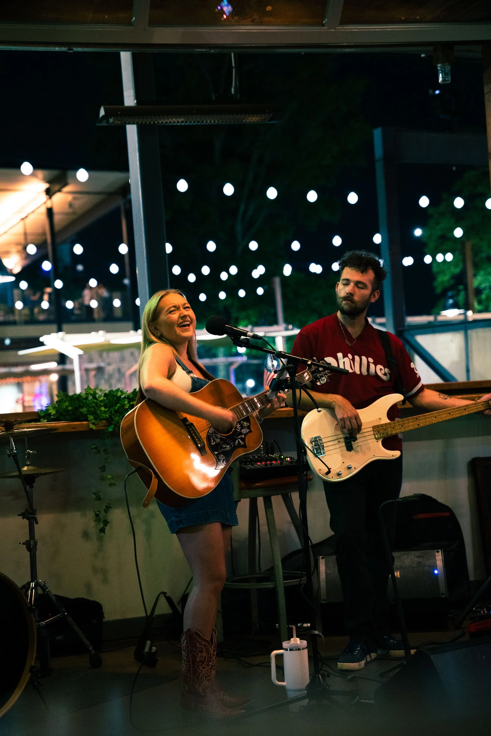 Two musicians performing at night under string lights outdoors. The woman is singing and playing an acoustic guitar, and the man is playing an electric bass guitar, both are standing and appear to be engaged in the music.