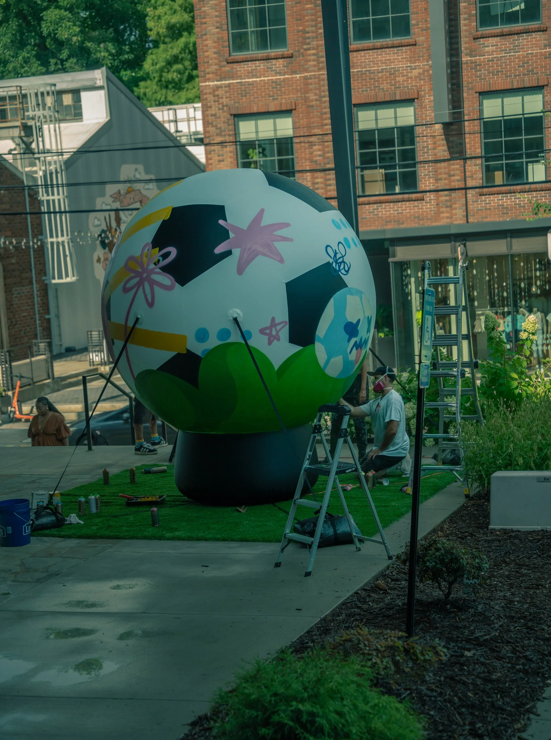 A large decorative sphere with colorful floral and cartoon-like patterns being assembled outdoors near a brick building and trees. There are ladders, tools, and people working on the display.