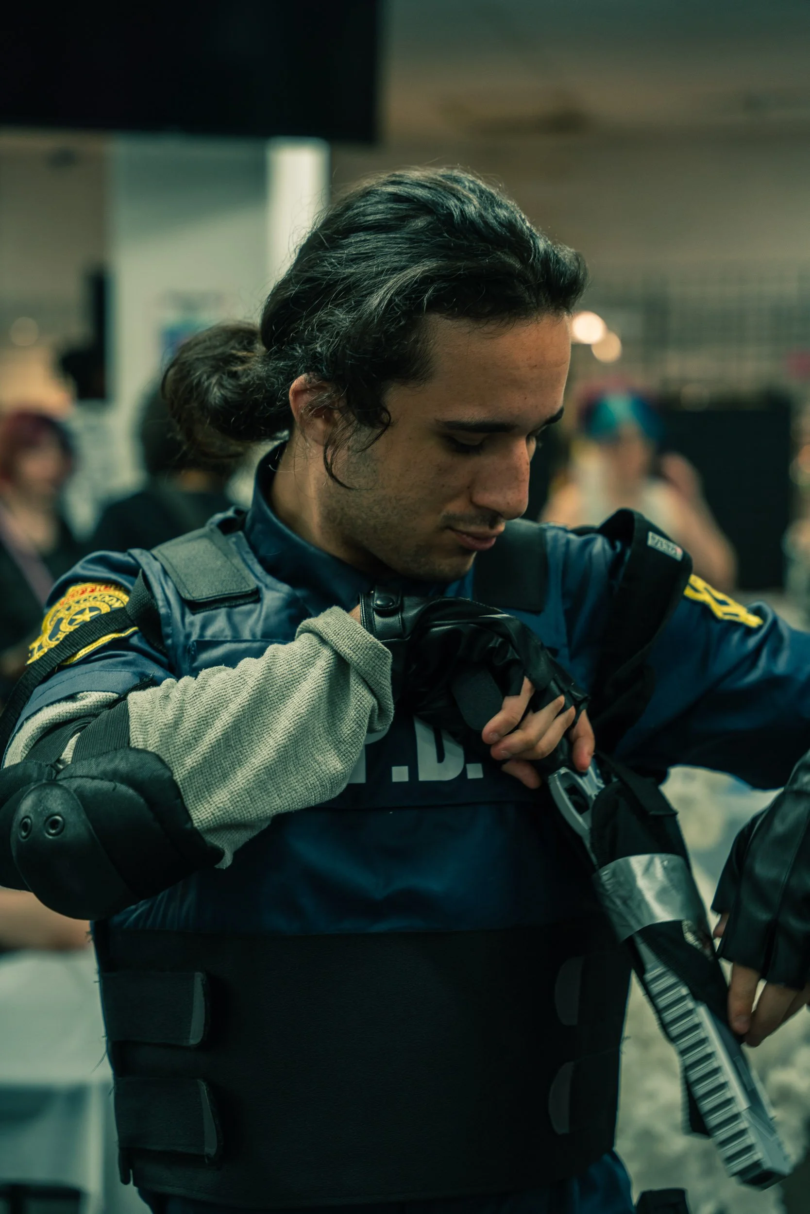 Close-up of a police officer with long dark hair tied back, adjusting a vest with a firearm in a crowded indoor setting.