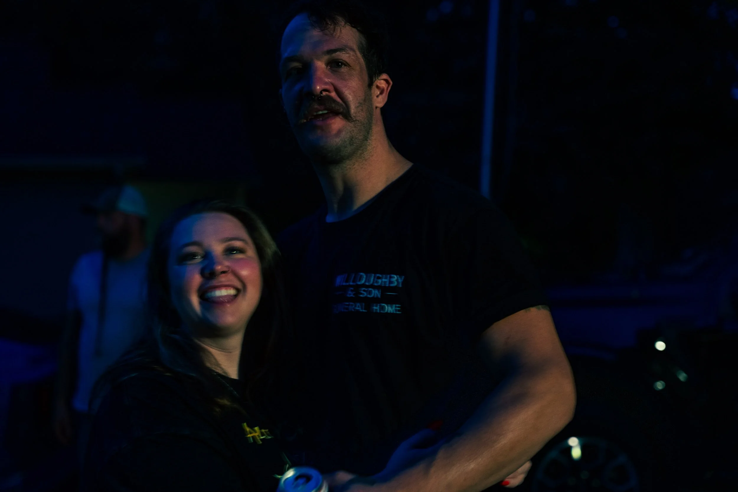 A smiling woman and a man with a mustache hugging at night, with the man wearing a black T-shirt and the woman having long hair.