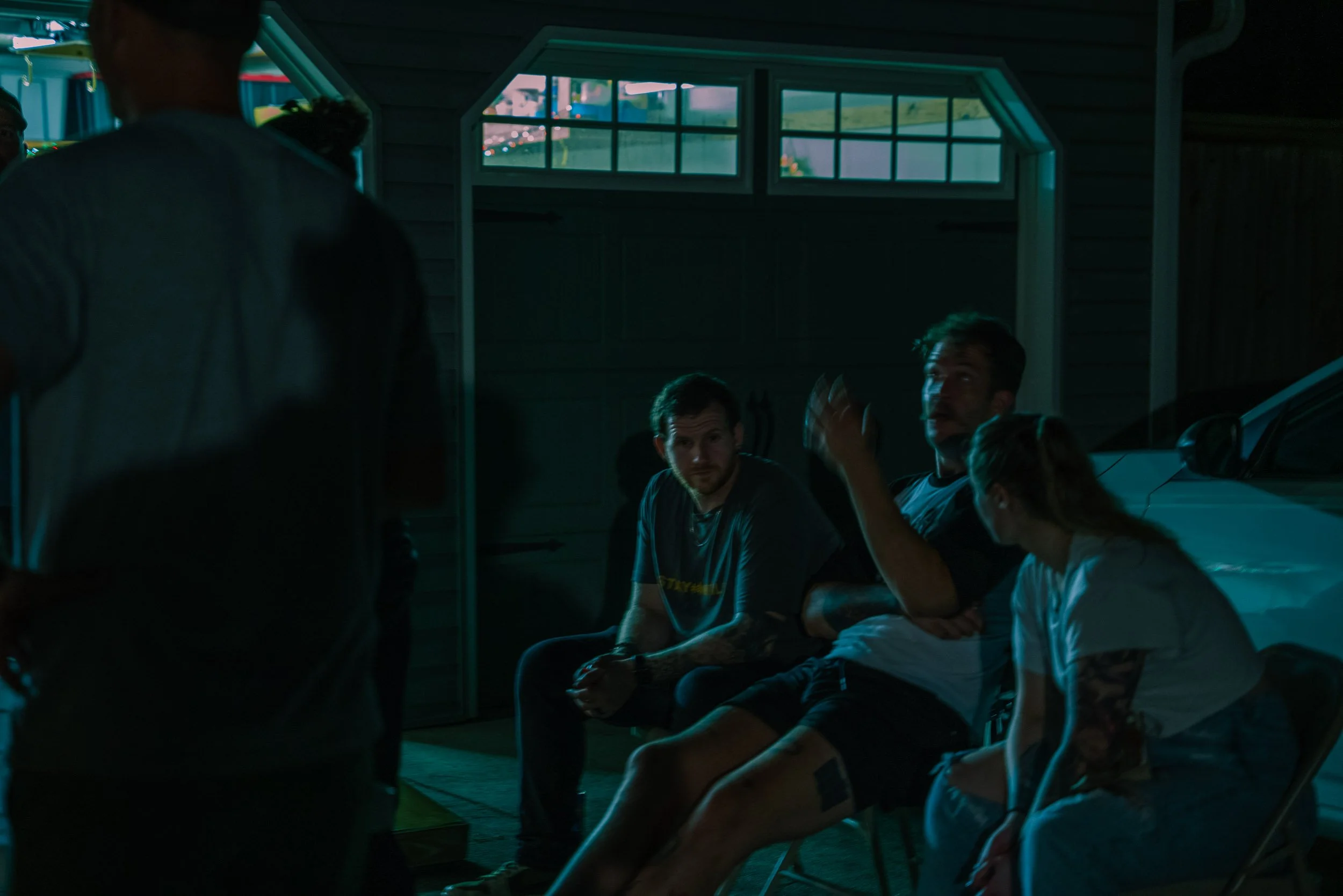 People sitting and engaging in conversation inside a dimly lit garage or workshop with a window showing a city street outside.
