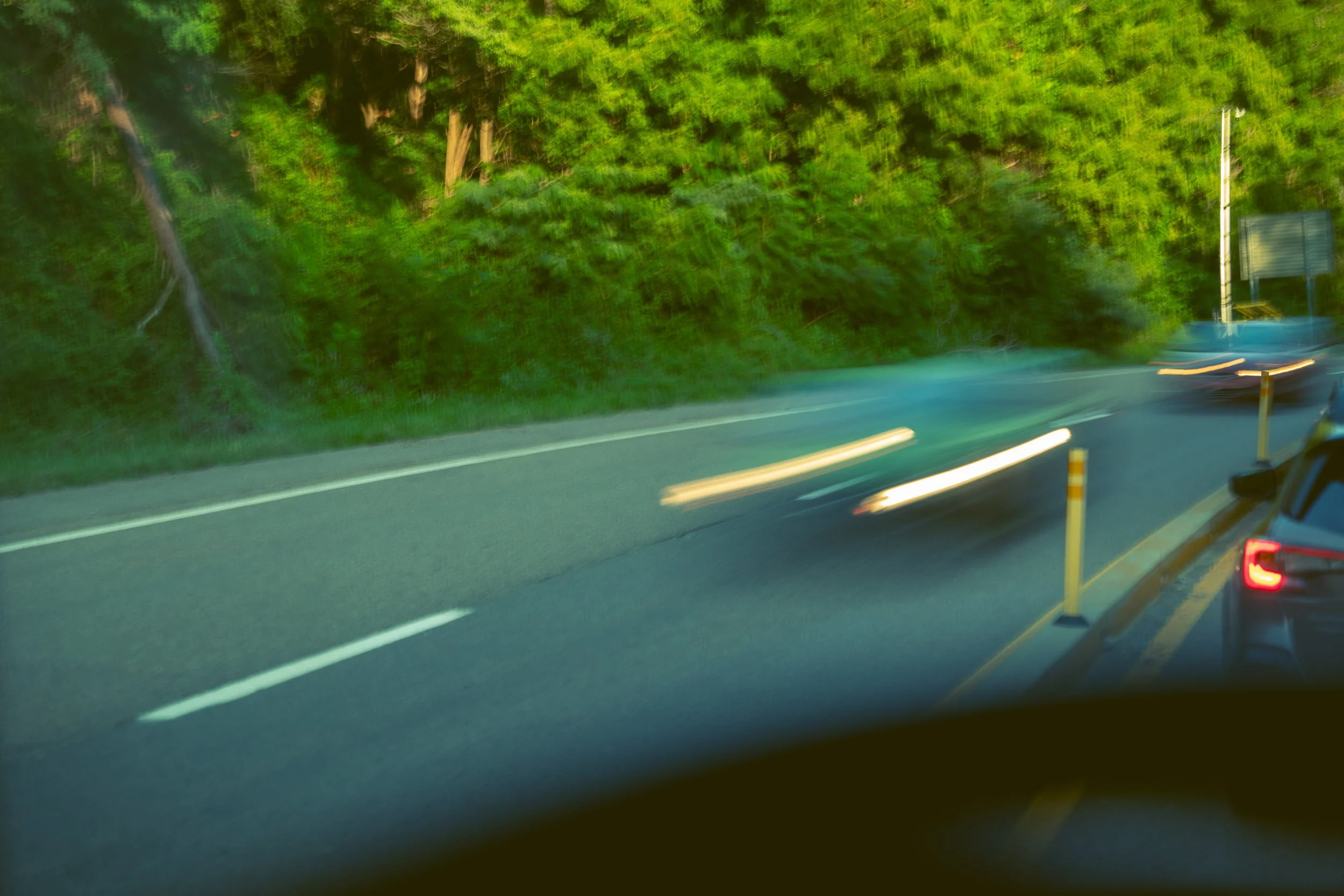 Photo of a highway with a blurred car passing by, green trees on the roadside, and roadside roadside signs.