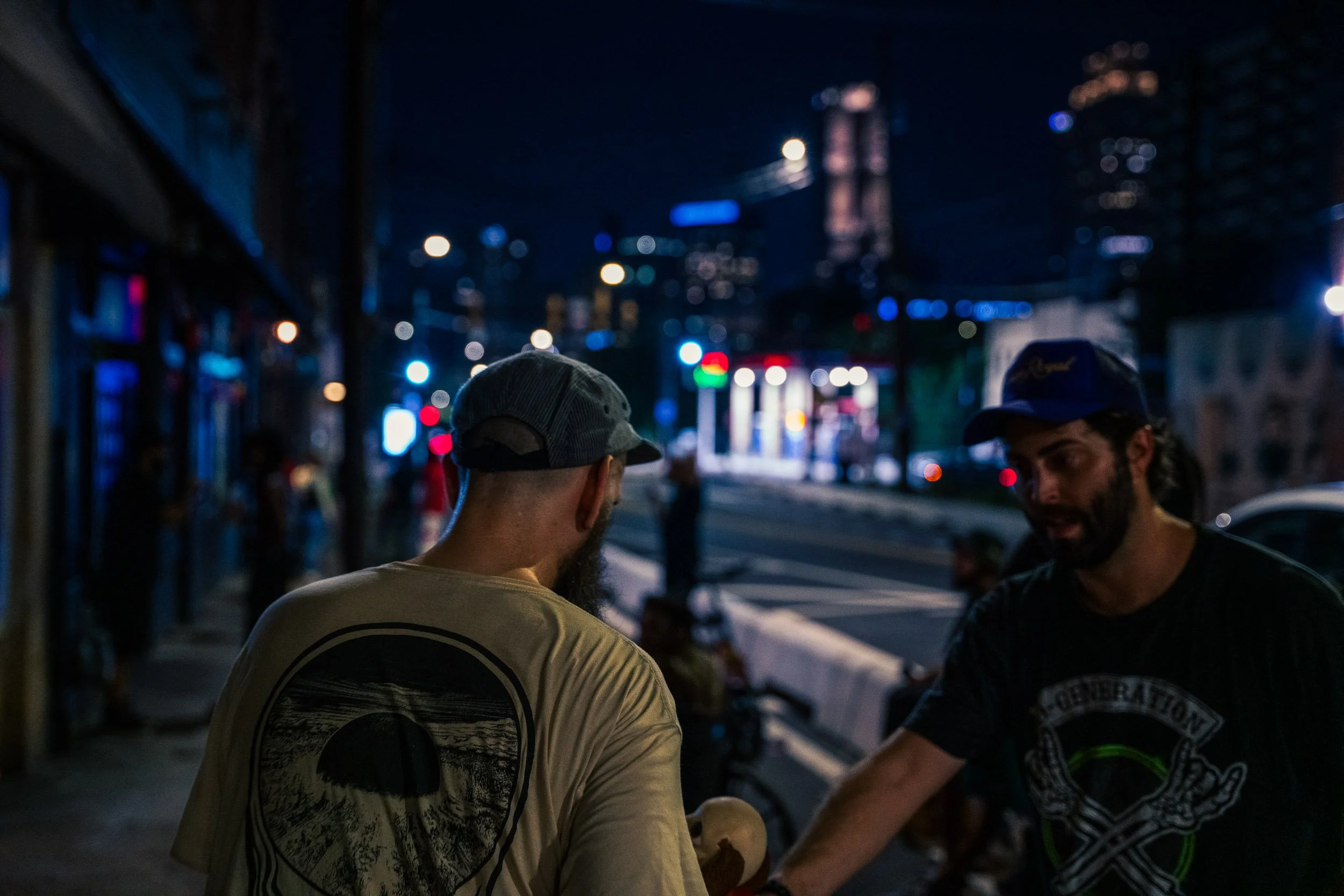 Two men with beards and hats stand on a city street at night, talking and smiling, with colorful blurred city lights and a tall building in the background.