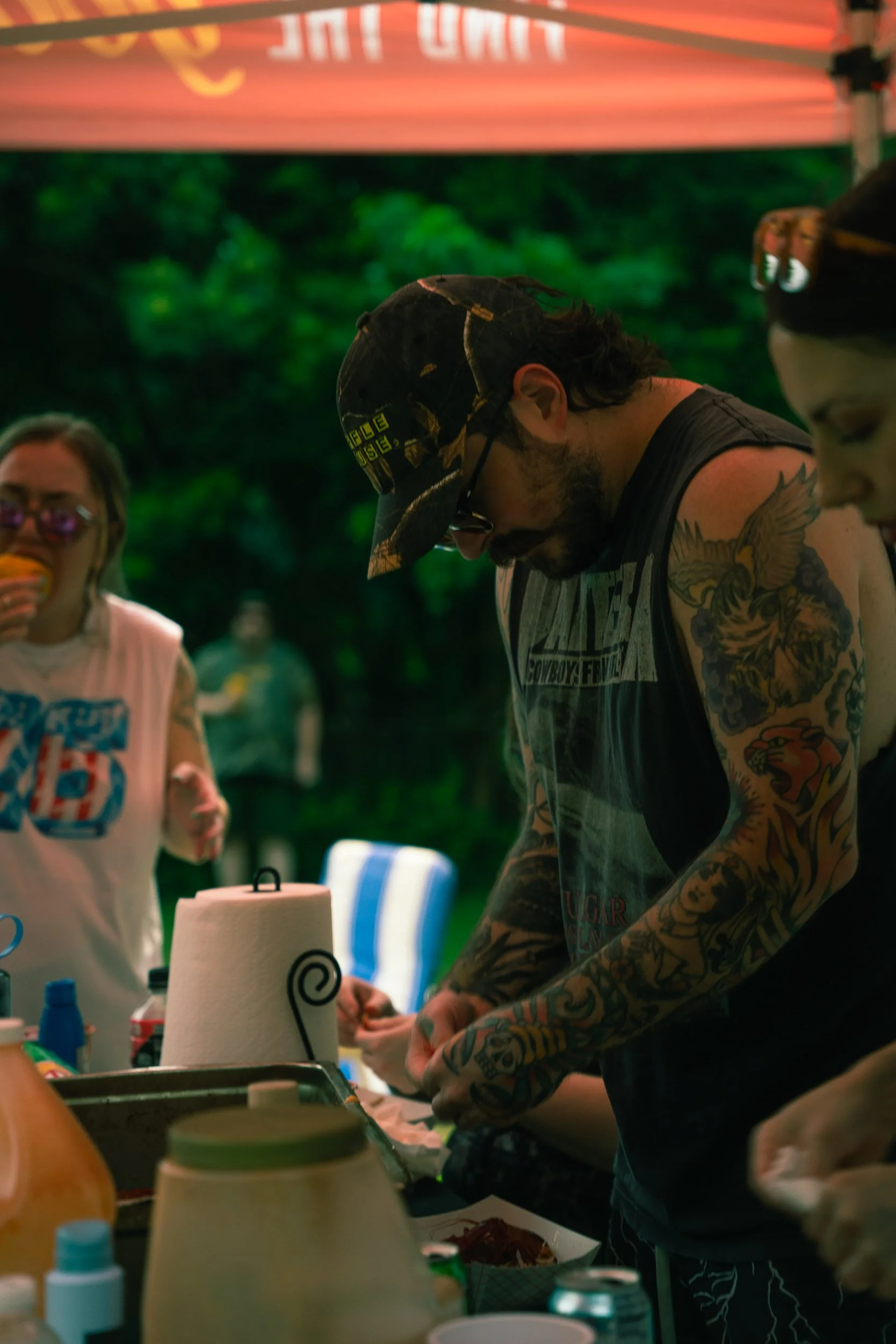 A man with tattoos, wearing glasses, a black hat, and a sleeveless shirt, preparing food at an outdoor gathering, with a woman in sunglasses and a white shirt in the background.