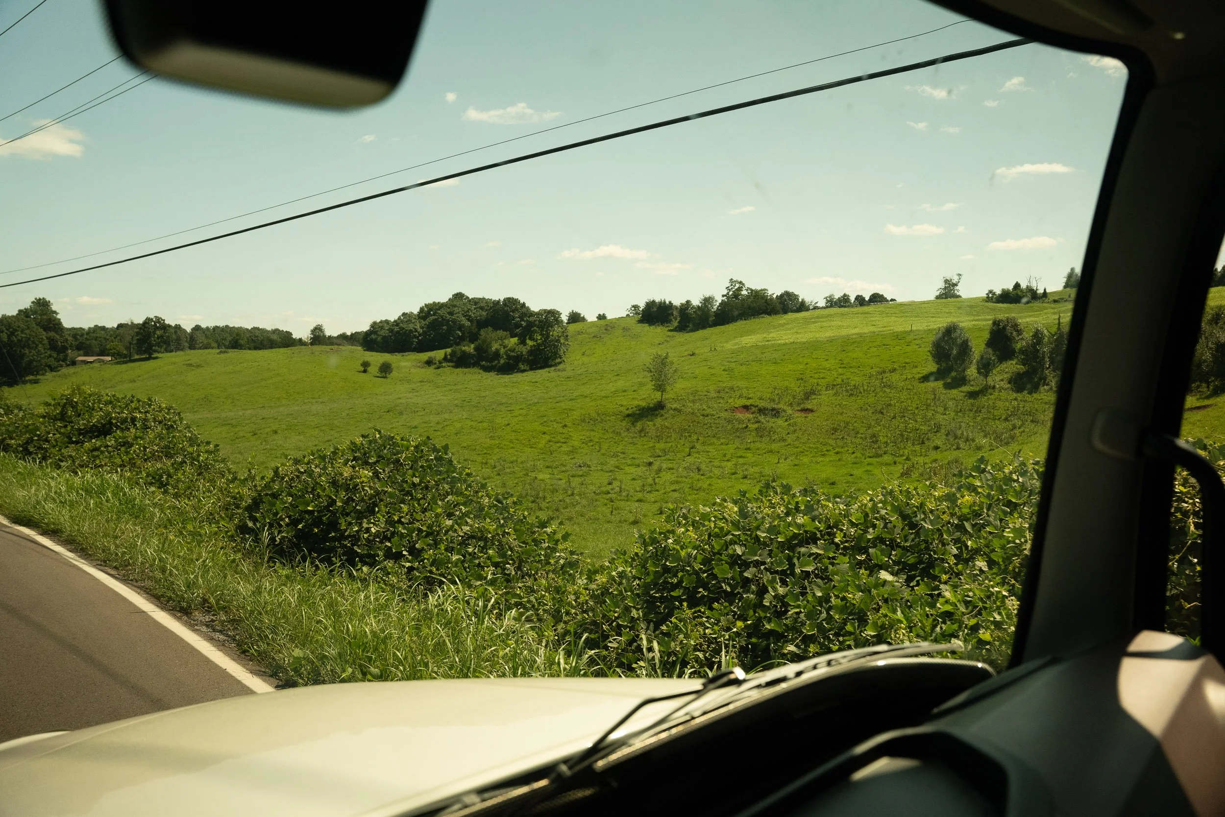 A view of green rolling hills and scattered trees seen through the windshield and side window of a vehicle, with power lines overhead and a clear sky with scattered clouds.