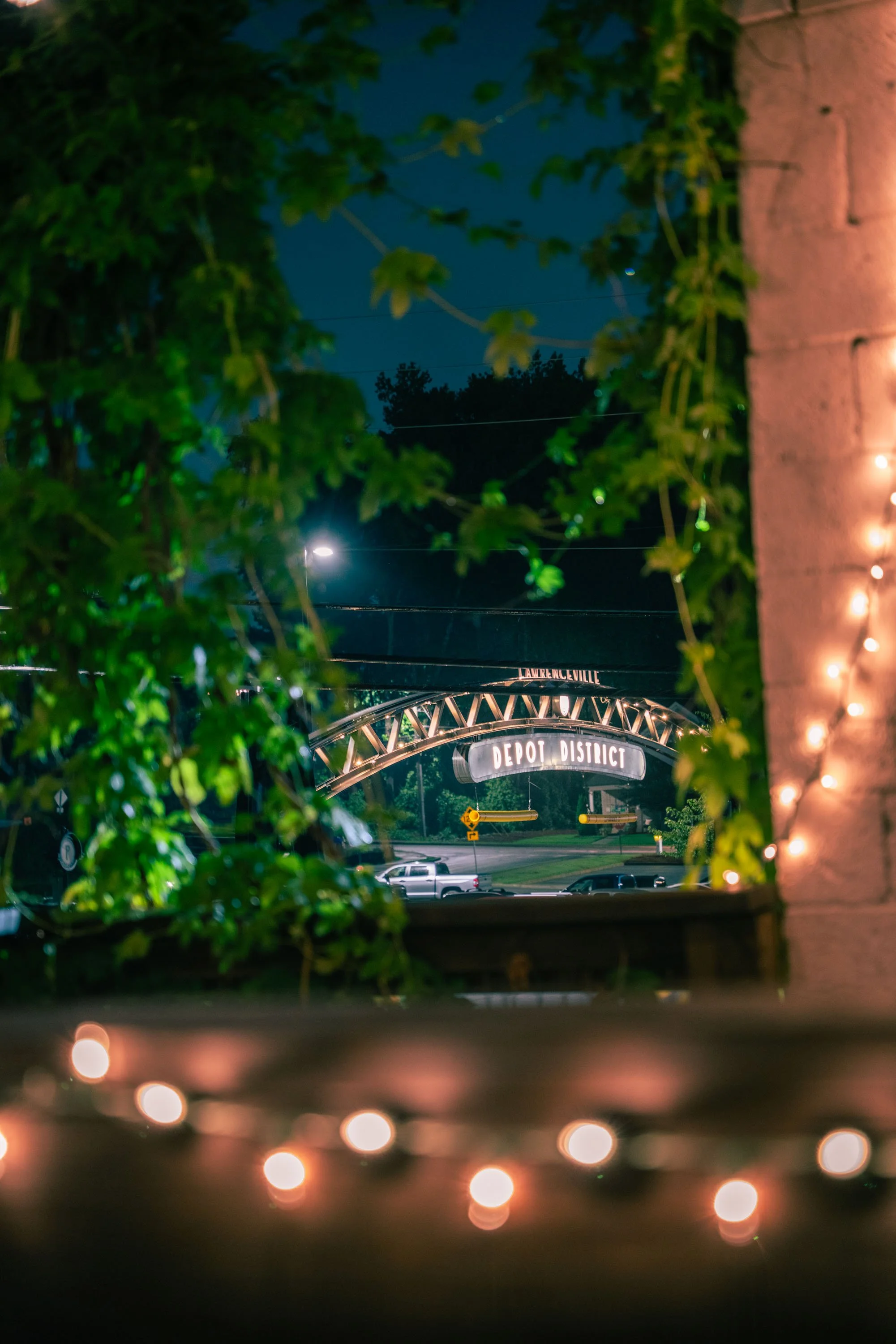 Nighttime view through a window with string lights, greenery on the sides, and a sign reading "Depot District" visible in the distance.