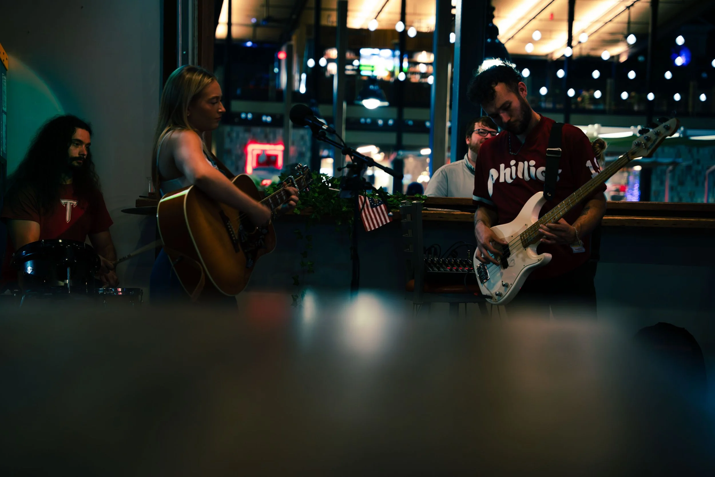 Four musicians perform indoors at night, with city lights visible through large windows behind them. One woman plays an acoustic guitar and sings into a microphone, while a man plays an electric bass guitar. The other two musicians, a man sitting behind a drum set and another man standing, listen and enjoy the performance.