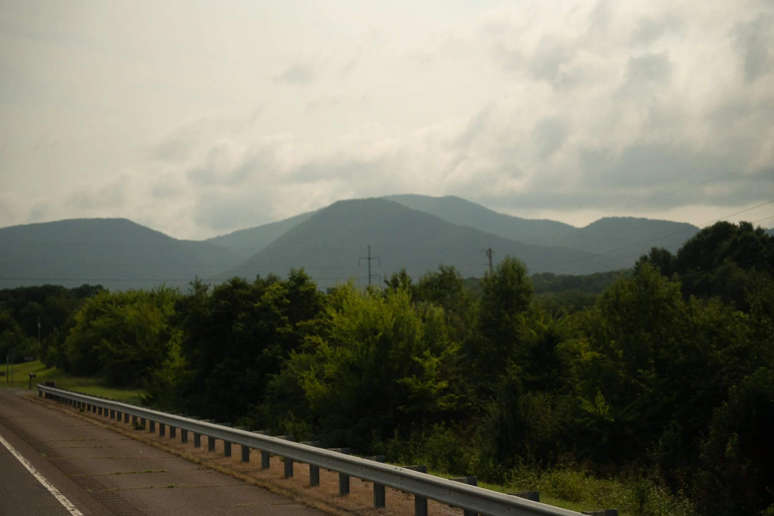 A scenic view of a mountainous landscape with cloudy sky, green trees, and power lines along the roadside.