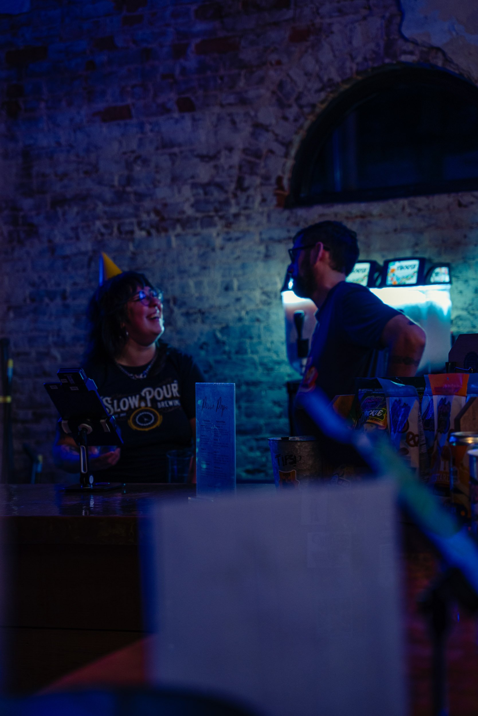 A woman and man are talking in a dimly lit bar or pub with a brick wall background. The woman is laughing and wearing a party hat, while the man is standing and wearing glasses, with a tattoo on his arm.