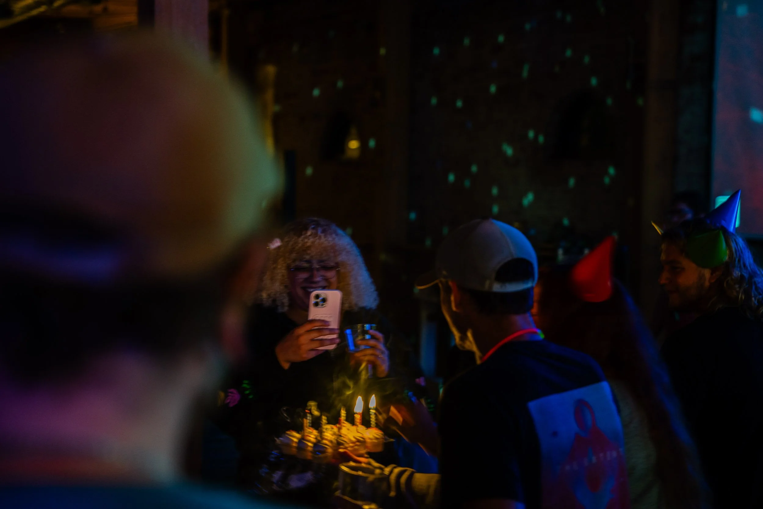 A group of people wearing party hats, gathered around a birthday cake with lit candles, in a dimly lit room with colorful dot patterns projected on the wall. One person is taking a photo with a smartphone.