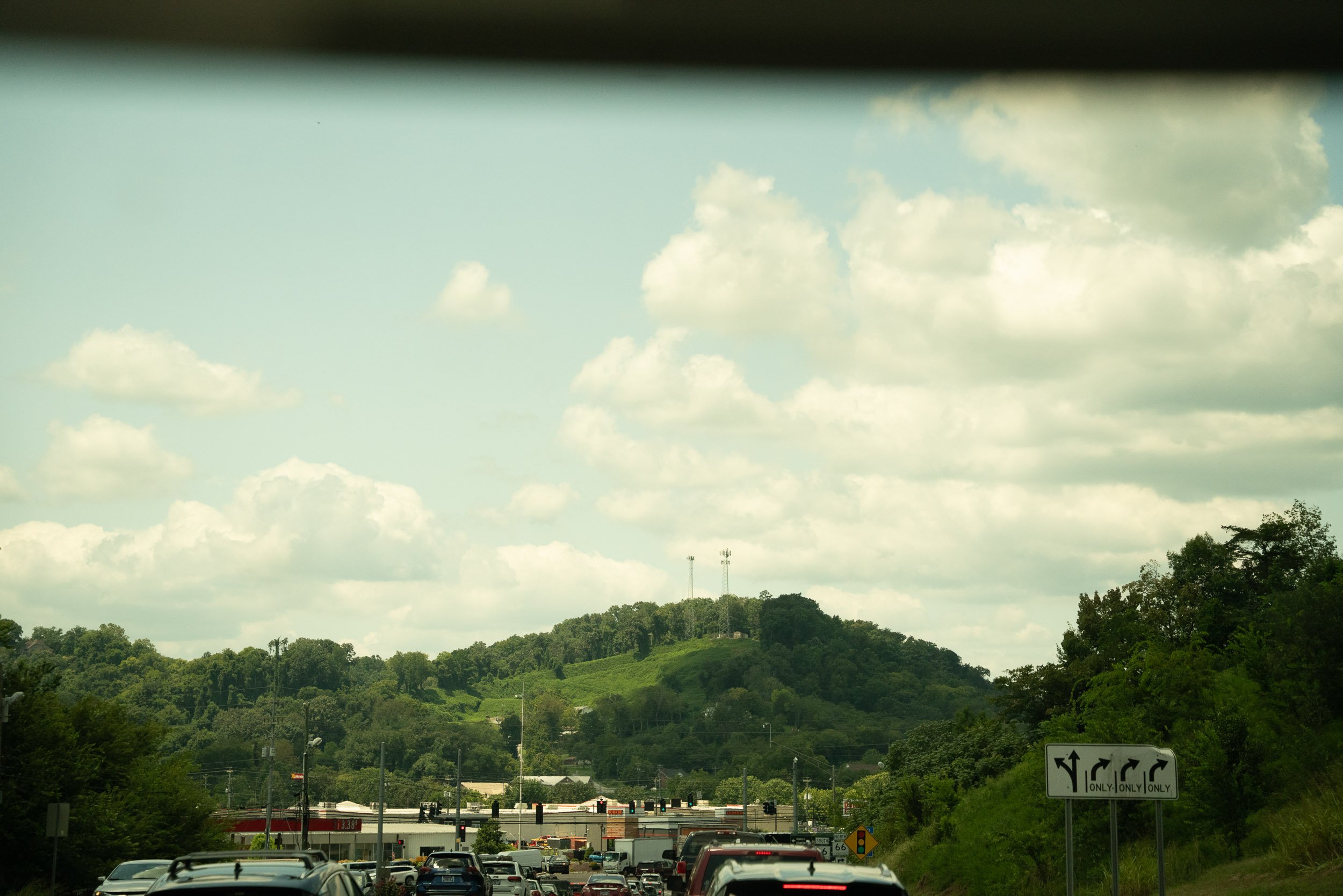 A busy highway with cars stopped at a traffic light, surrounded by lush green hills and trees, under a partly cloudy sky.