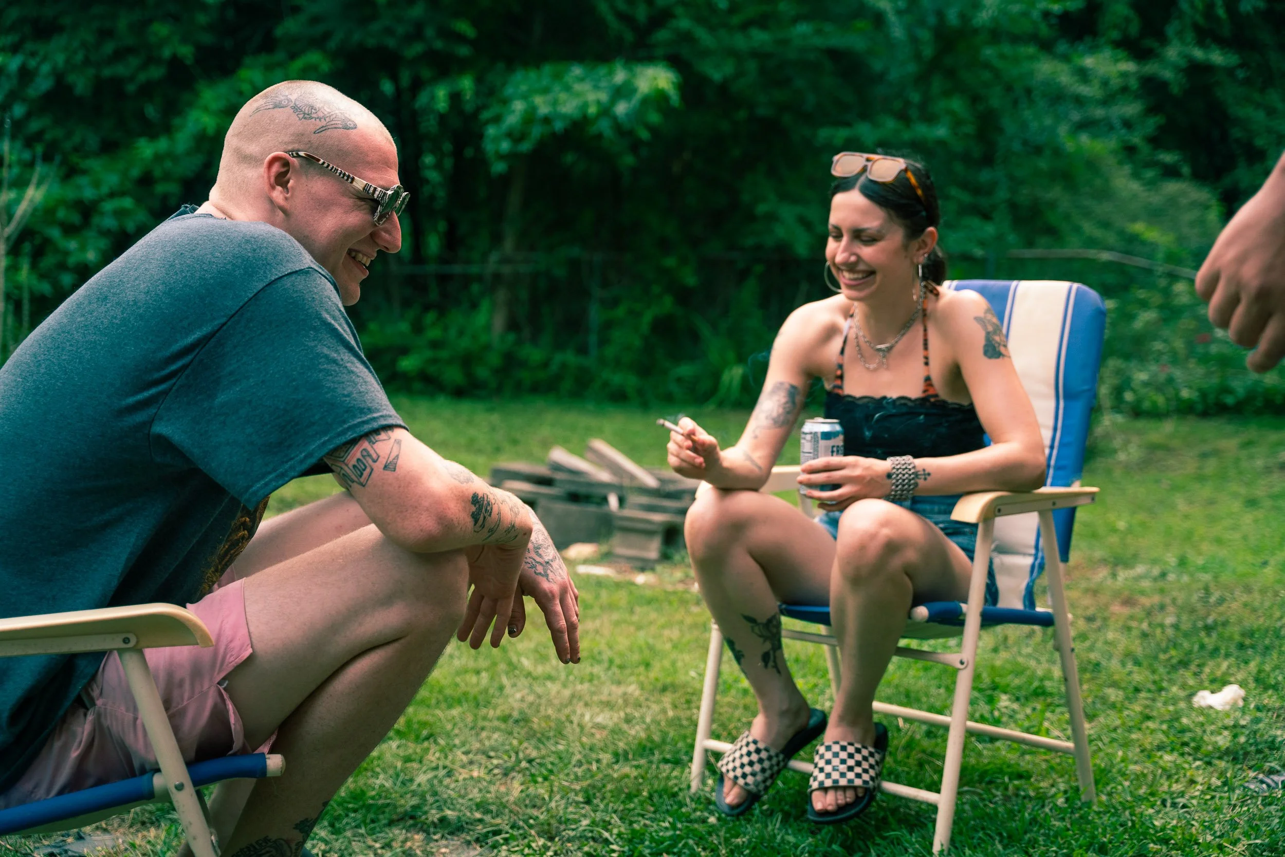 Two people sitting outdoors on lawn chairs, smiling and laughing. One is a bald man with sunglasses, tattoos, and a grey t-shirt, the other a woman with short black hair, sunglasses on her head, and tattoos, wearing a black top. They appear to be enj