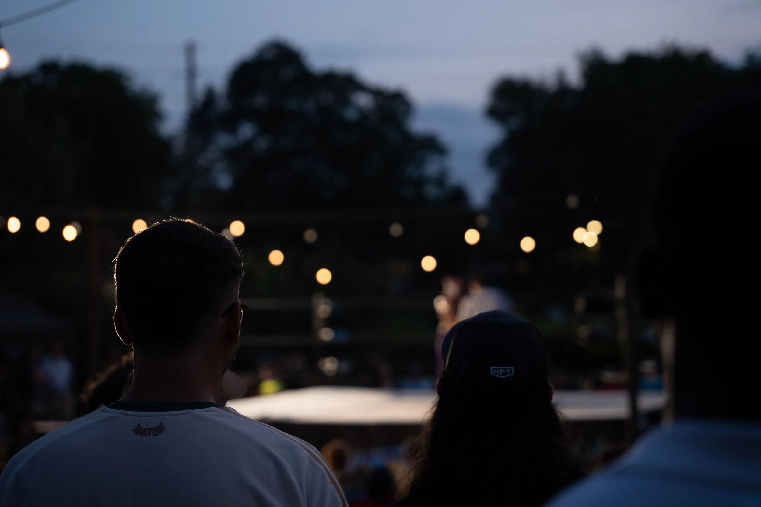 People watching an outdoor movie at dusk with string lights and trees in the background.