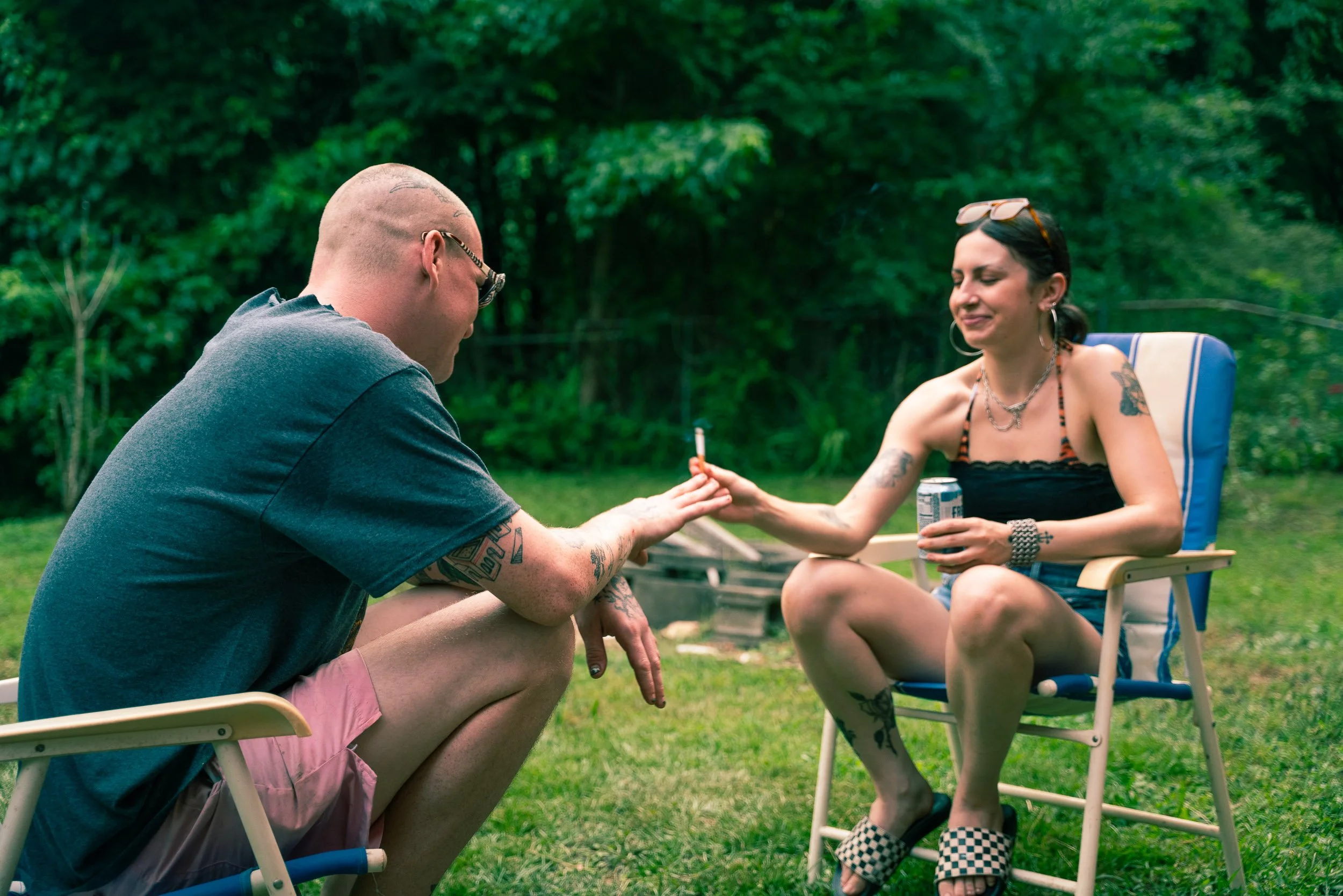 A man and woman sitting outside on lawn chairs, engaging in a handshake or hand gesture, with trees and grass in the background.