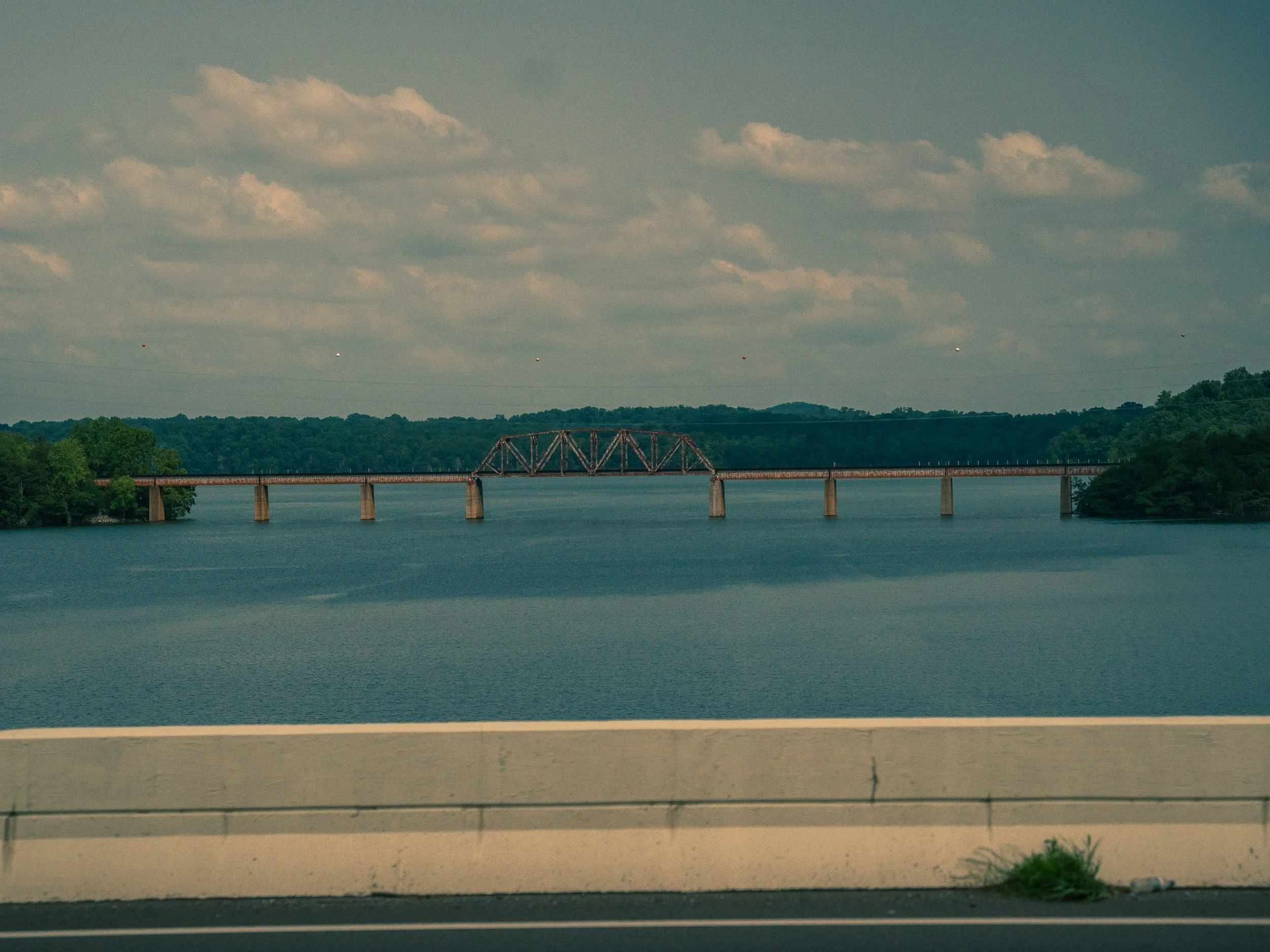 Bridge over a wide river or lake with green trees on both banks and a sky with scattered clouds in the background.