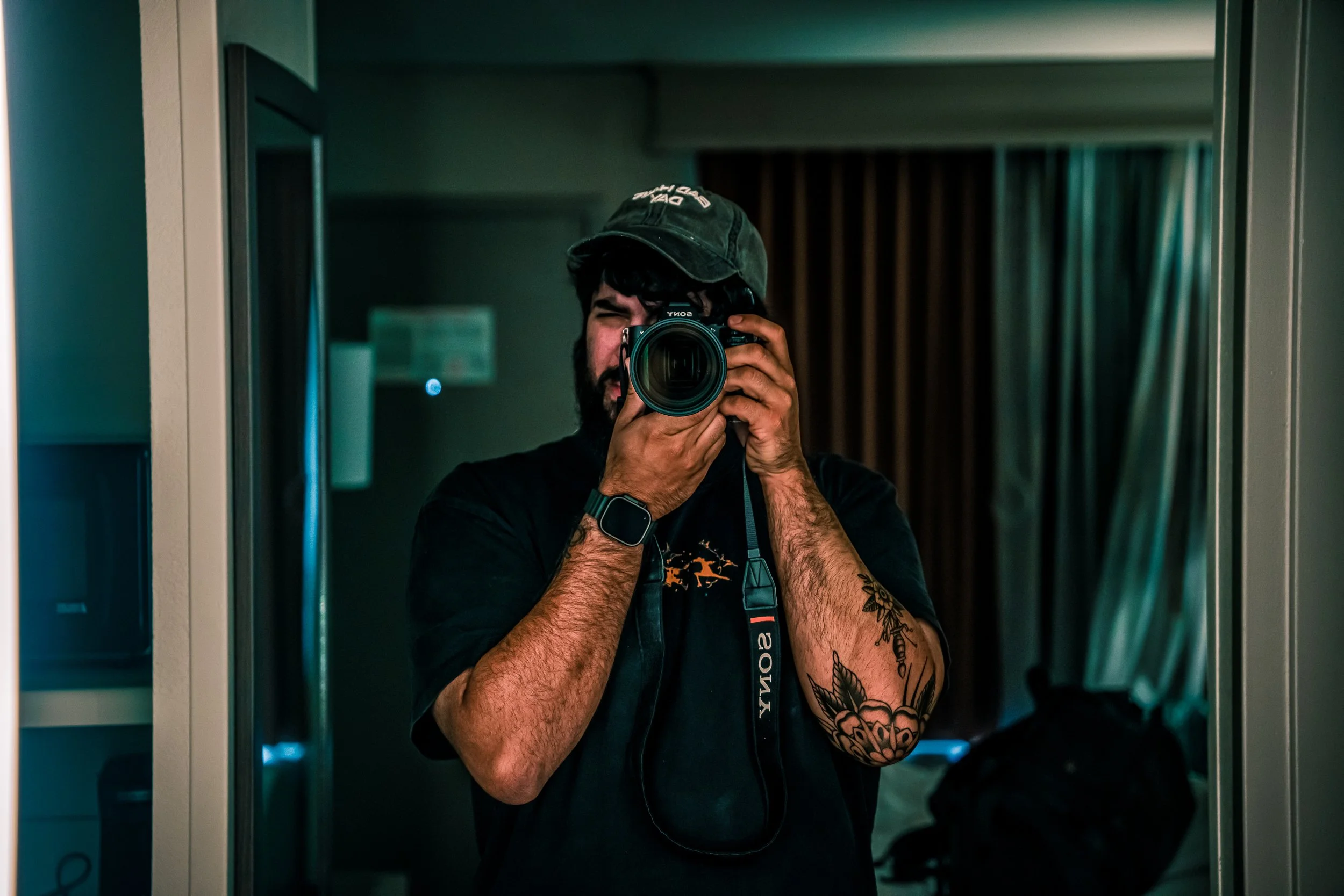 Person taking a photo in a mirror, wearing a black T-shirt, a green cap, holding a camera, and has tattoos on his left arm.