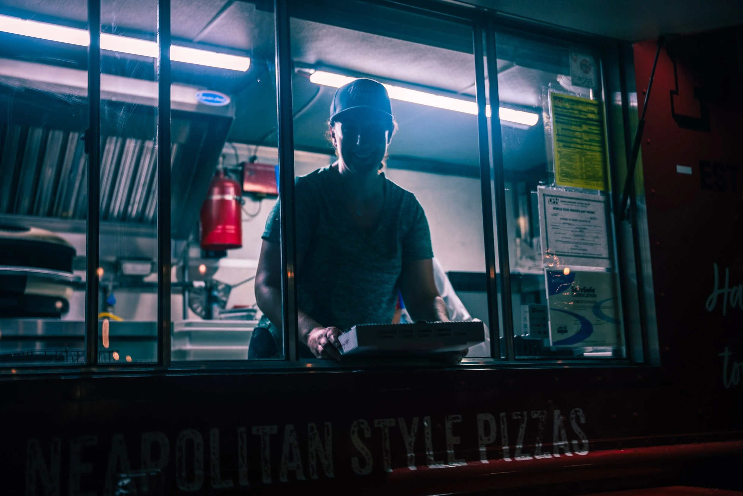 A person wearing a baseball cap and glasses is working inside a food truck, seen through a window with metal bars. The person appears to be handling some equipment or preparing food, with bright fluorescent lights overhead and various kitchen items v