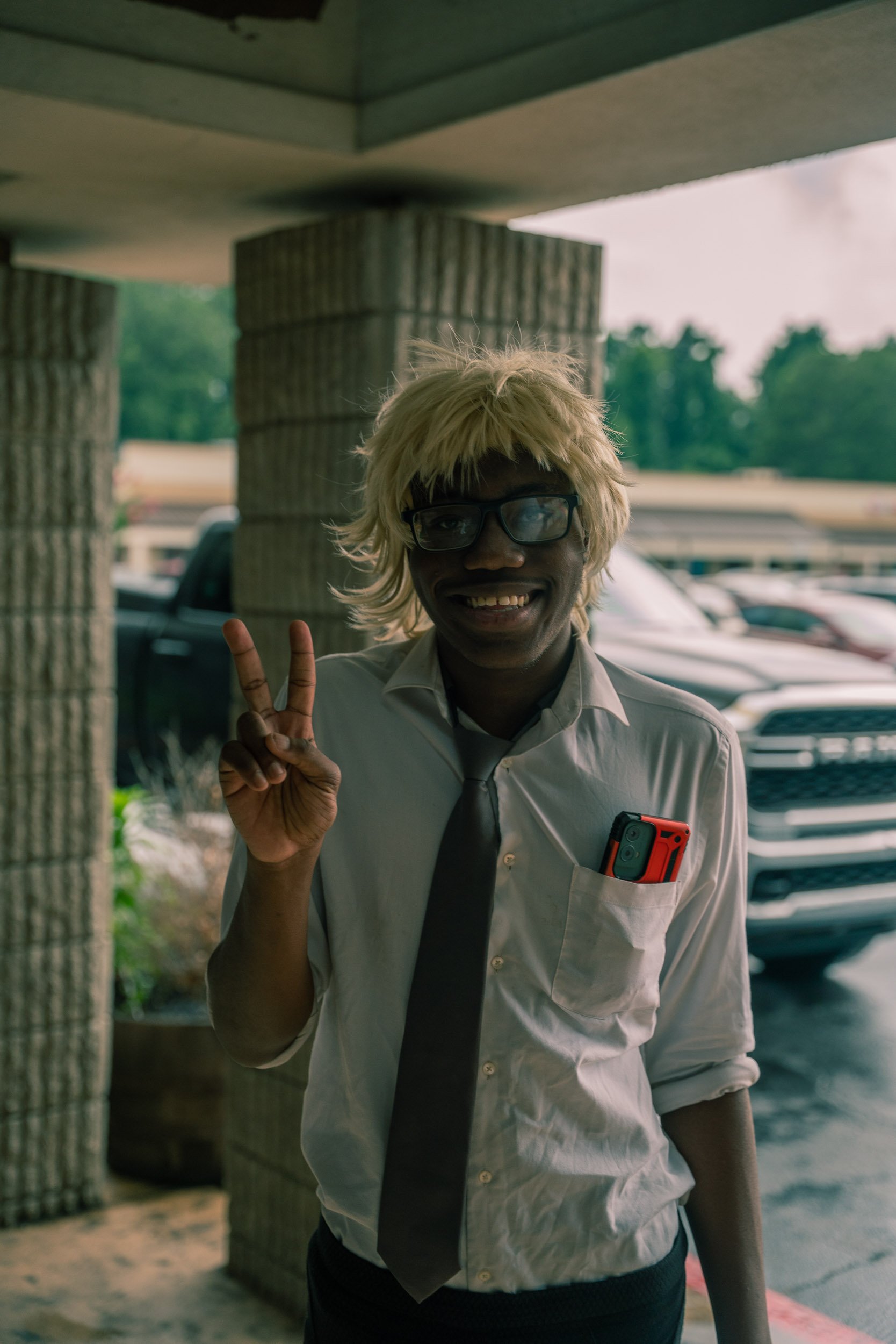 Person with blond hair, glasses, white shirt, and black tie smiling and making a peace sign in an outdoor parking lot.
