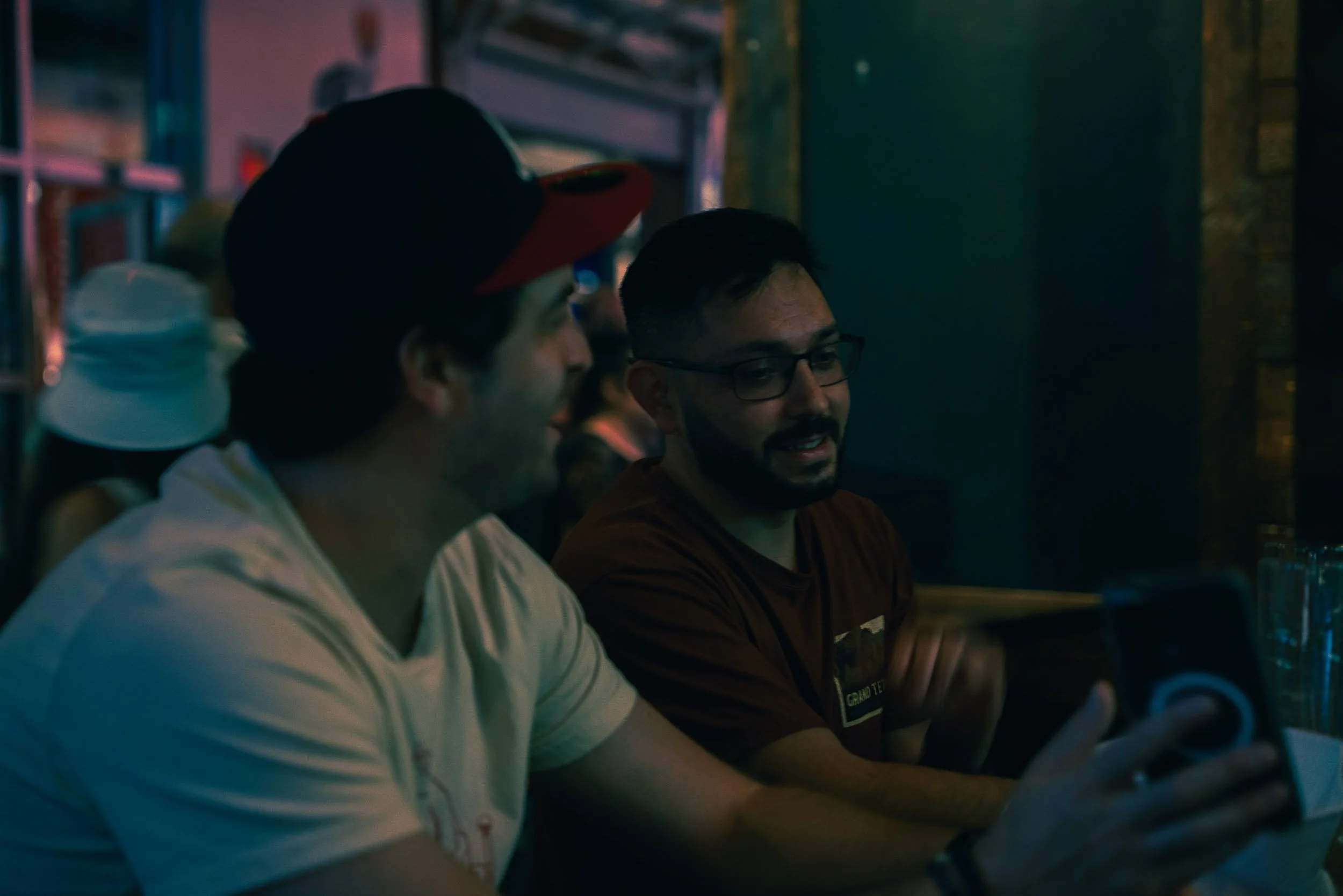 Two men sitting at a bar, one wearing a white shirt and the other wearing glasses and a brown shirt, engaged in conversation in a dimly lit setting.