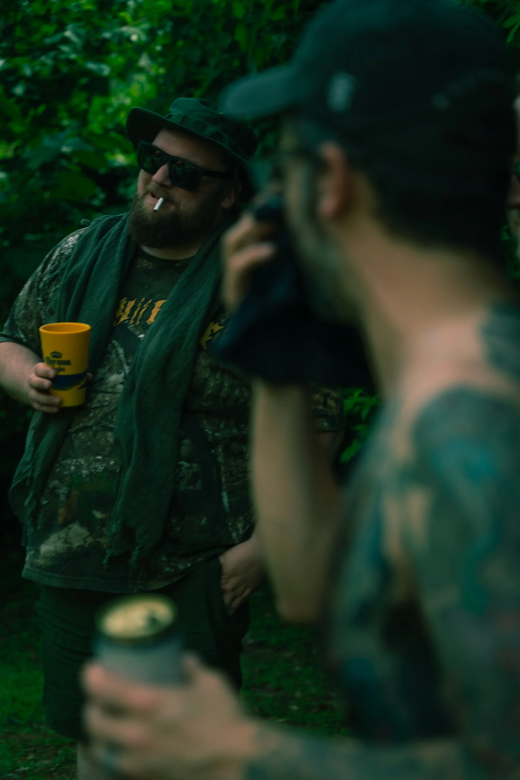 A man with a beard wearing sunglasses, a camouflage hat, a camouflage shirt, and a dark jacket, holding a yellow cup, talking to a person in dark sunglasses and a jacket, in a forest setting.