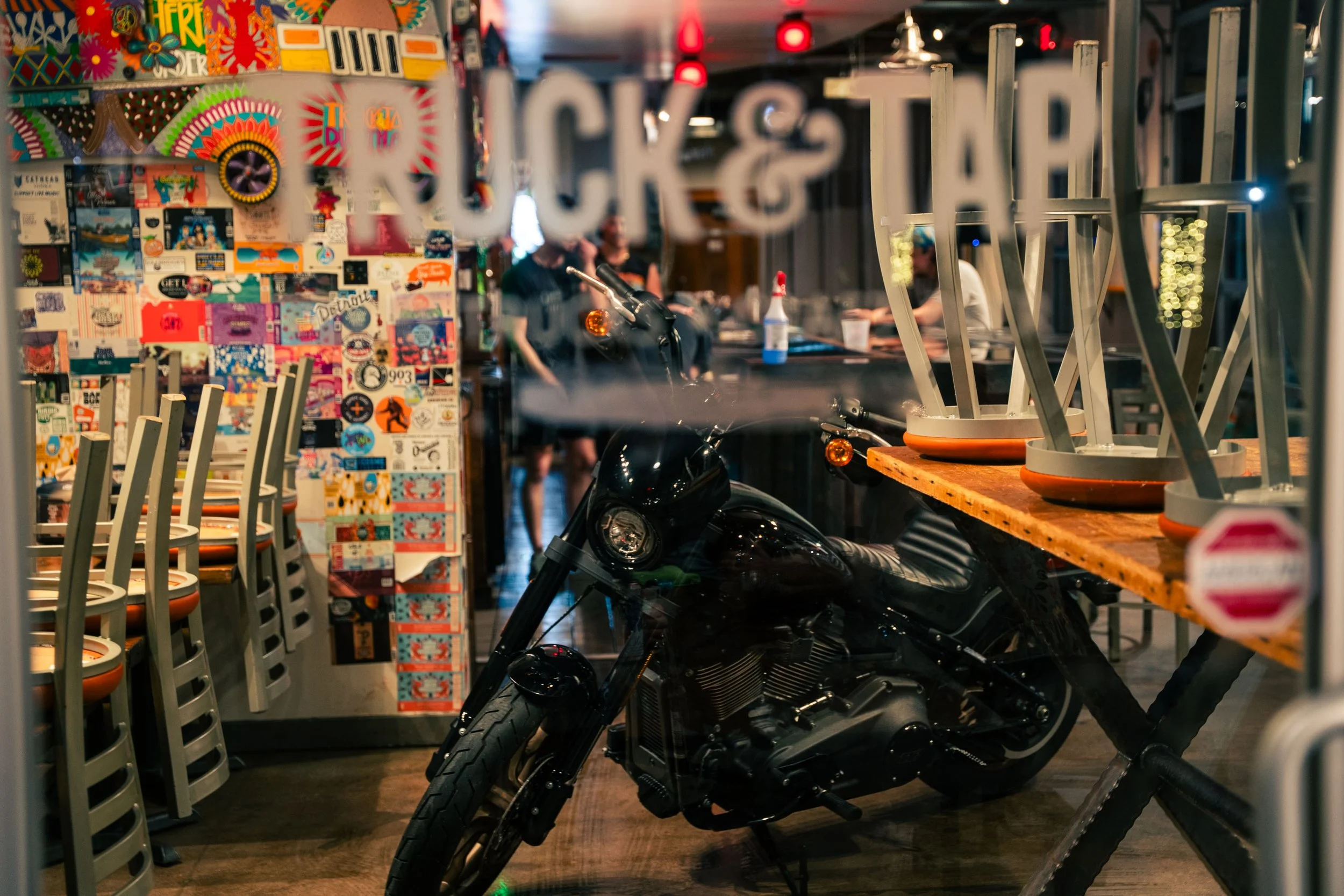 A motorcycle parked inside a restaurant, viewed through a glass window with white text. In the background, people are sitting at a bar or table, with colorful posters and decorated walls, and stacked chairs and tables.