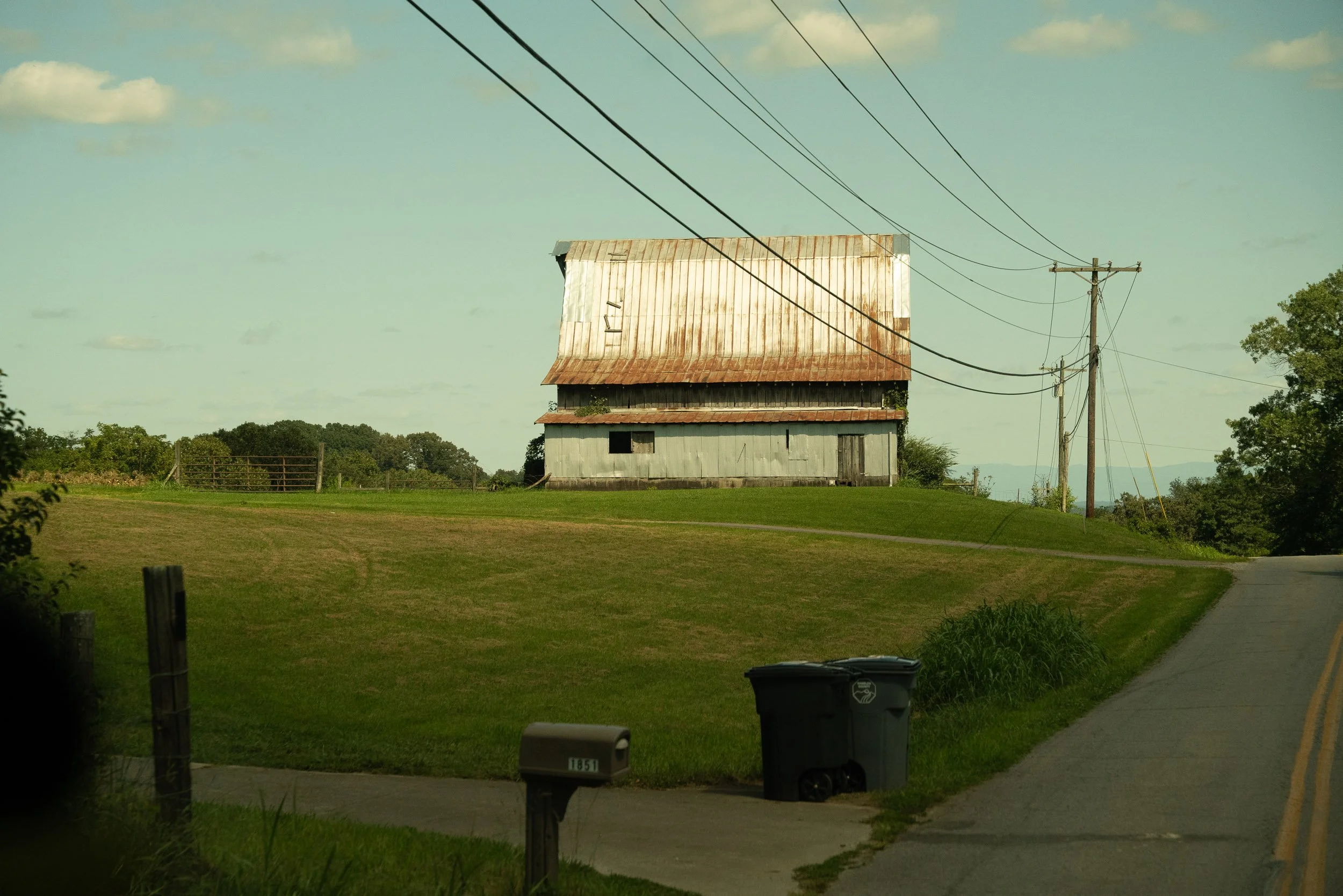 A rural scene with a rusty metal barn and power lines against a clear sky with scattered clouds.