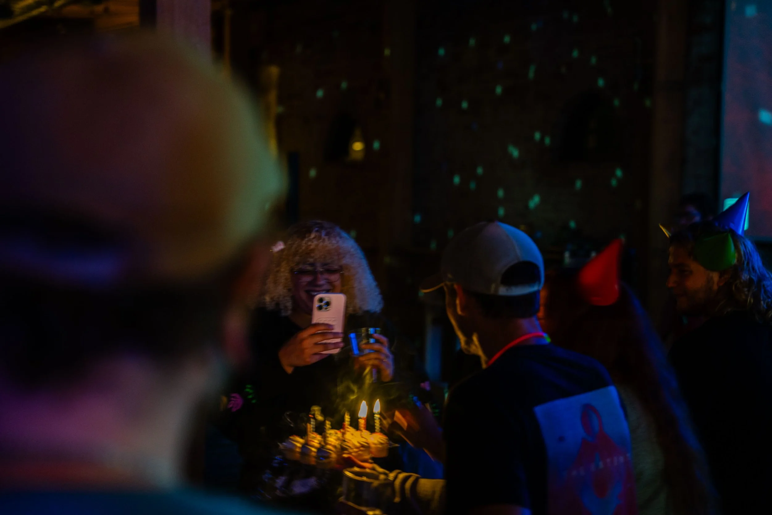 People at a celebration or birthday party in a dimly lit room with colorful party hats, a birthday cake with lit candles, and a woman taking a photo.