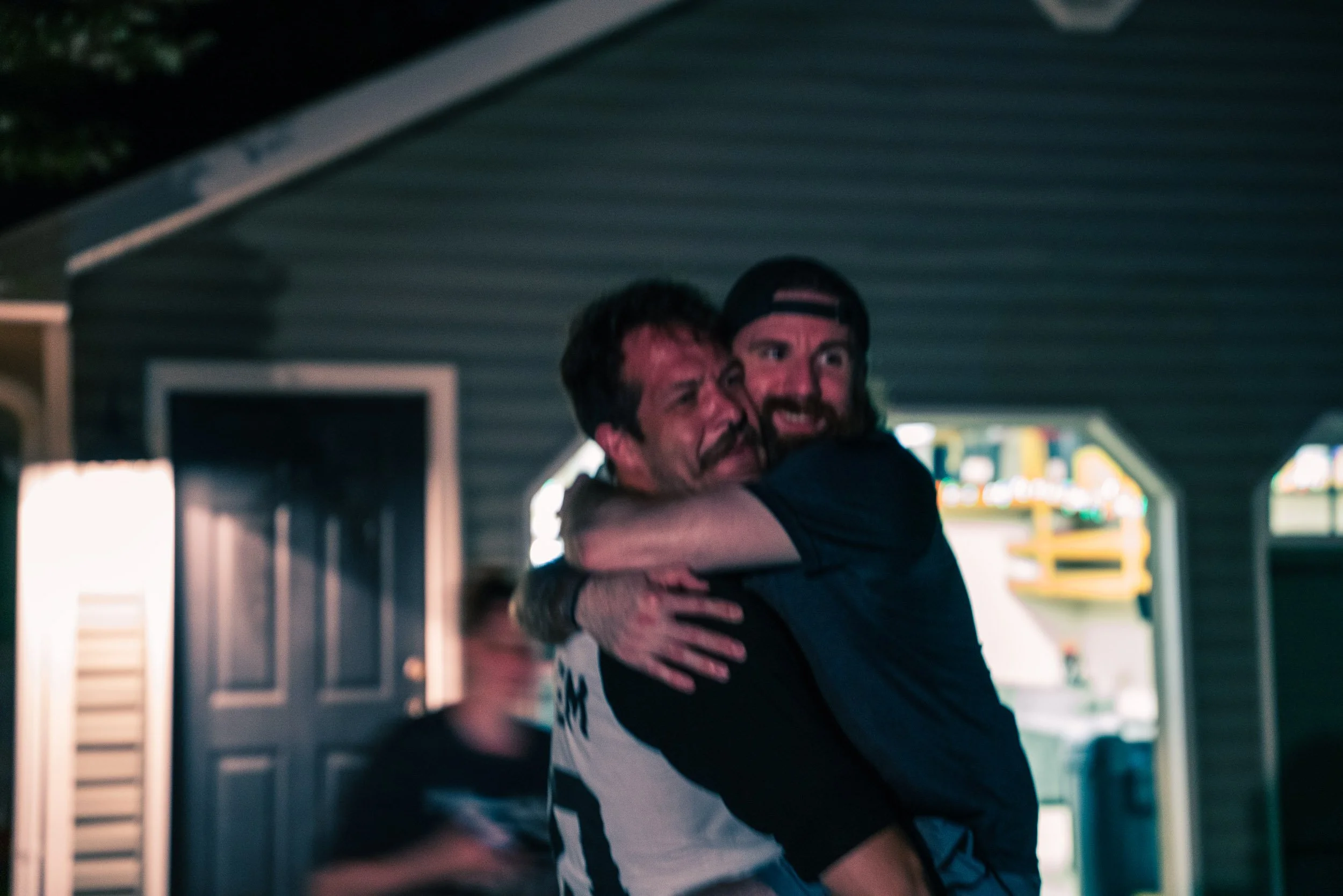 Two men hugging inside an arcade or entertainment venue, with neon and bright lights in the background.